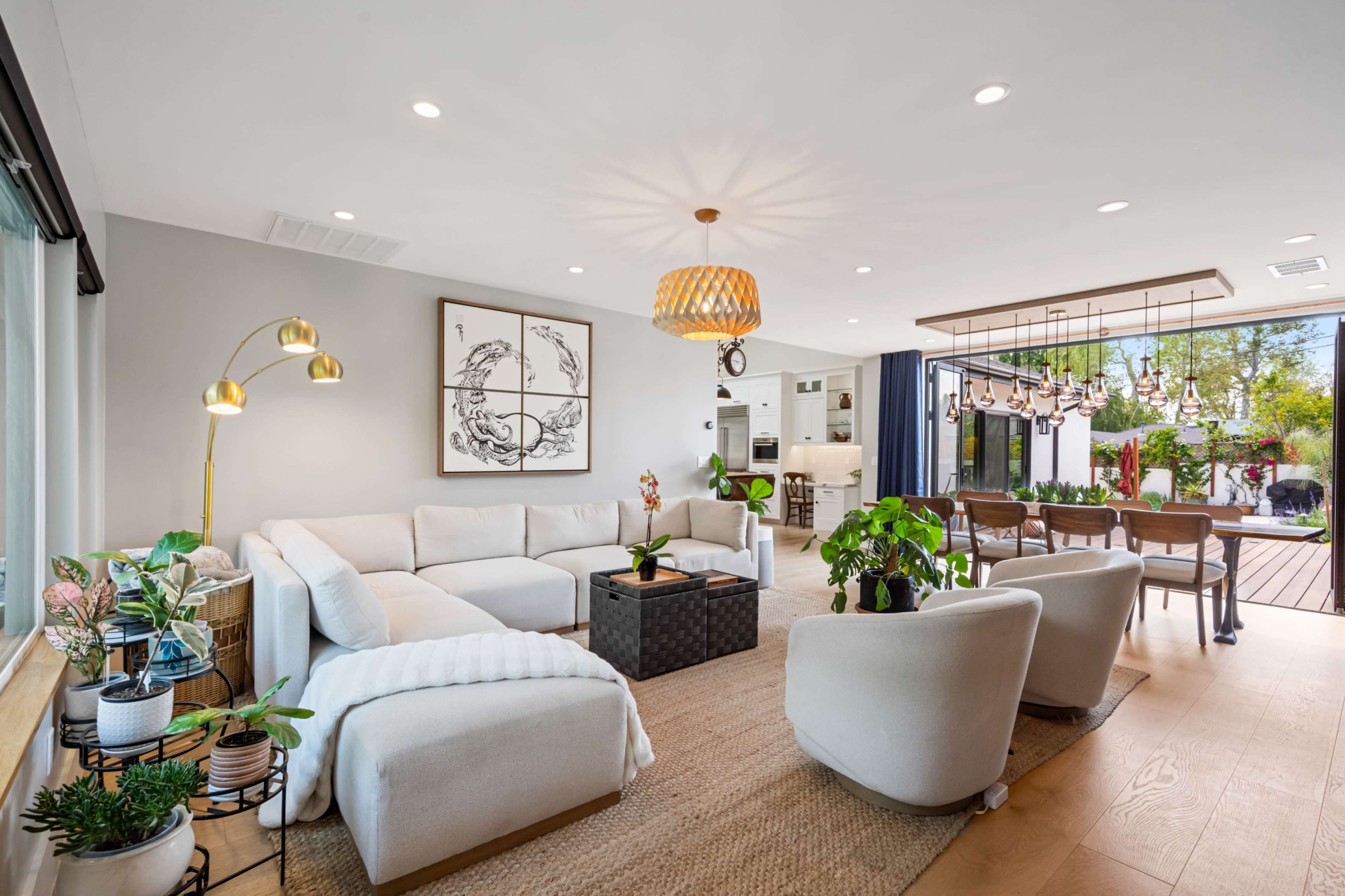 The image shows a modern, open-concept living room with a large white sectional sofa, a coffee table, plants, and a view into a dining area featuring large windows and a patio beyond.