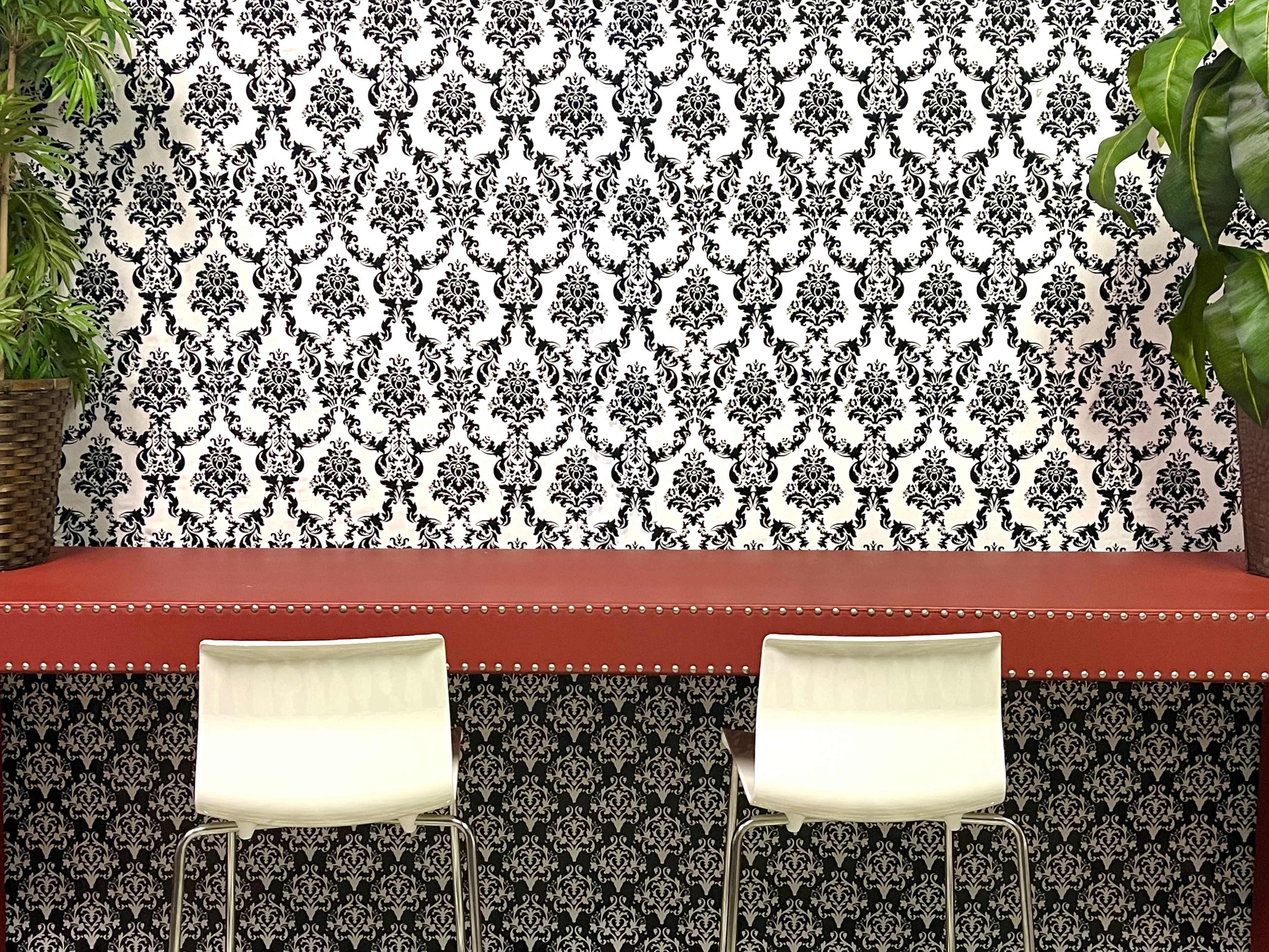 A red countertop is set against a black and white damask-patterned wall, flanked by two white chairs and potted plants.