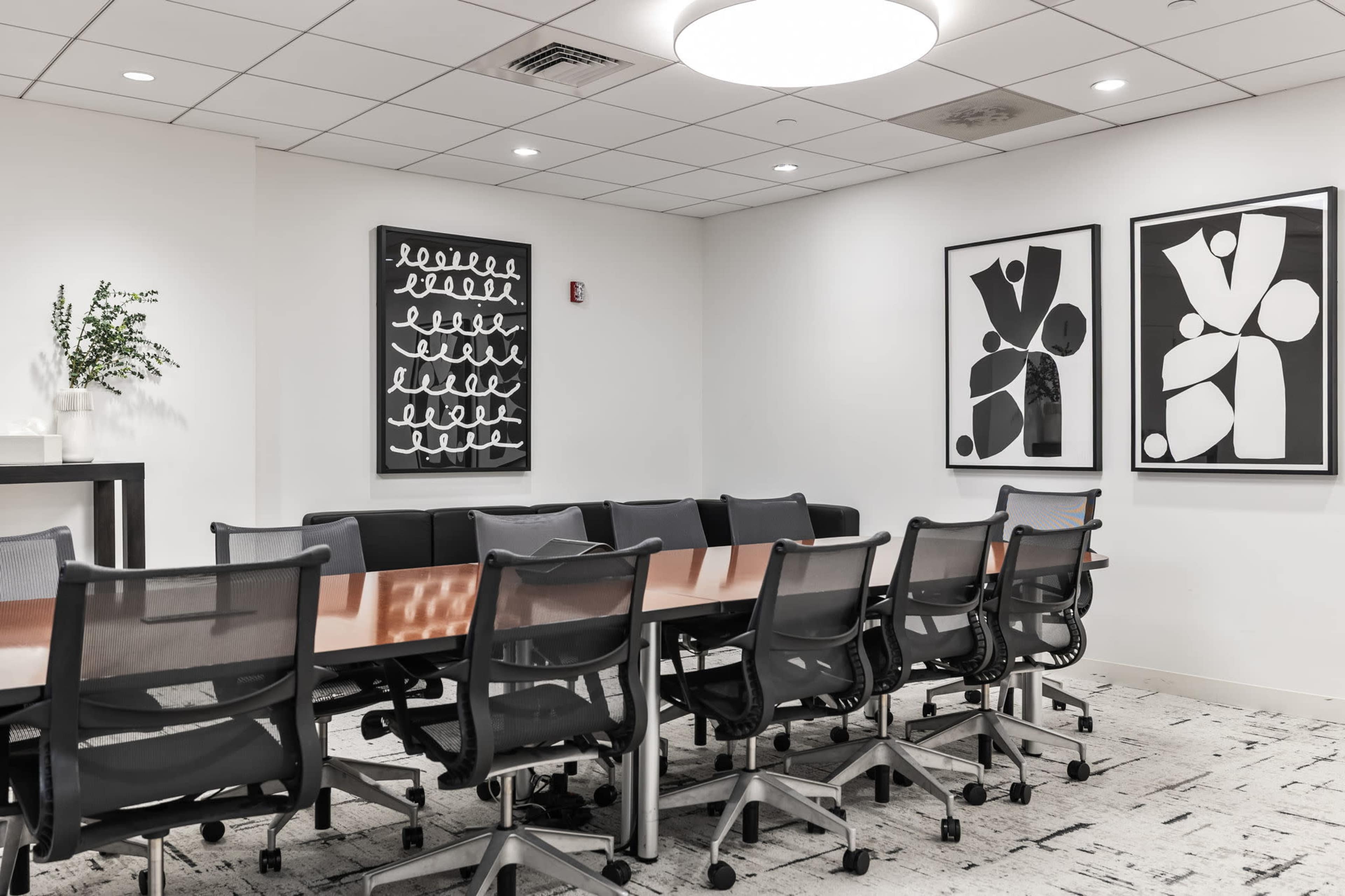 The image shows a modern conference room with a rectangular table surrounded by chairs, featuring abstract black and white artwork on the walls.