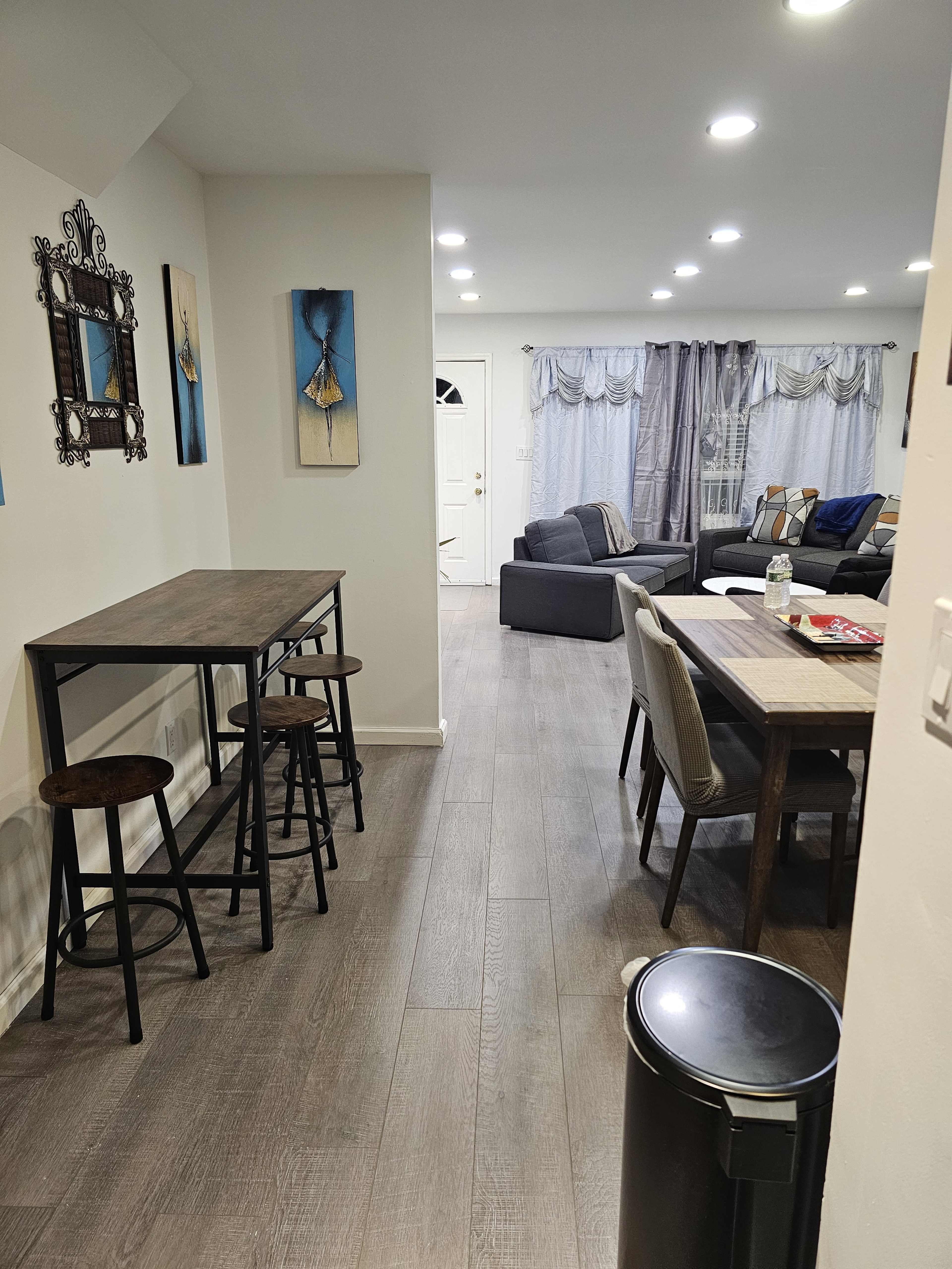A modern, open-concept living space that includes a dining area with a table and chairs, a tall table with stools, and a seating area with a sofa and curtains in the background.