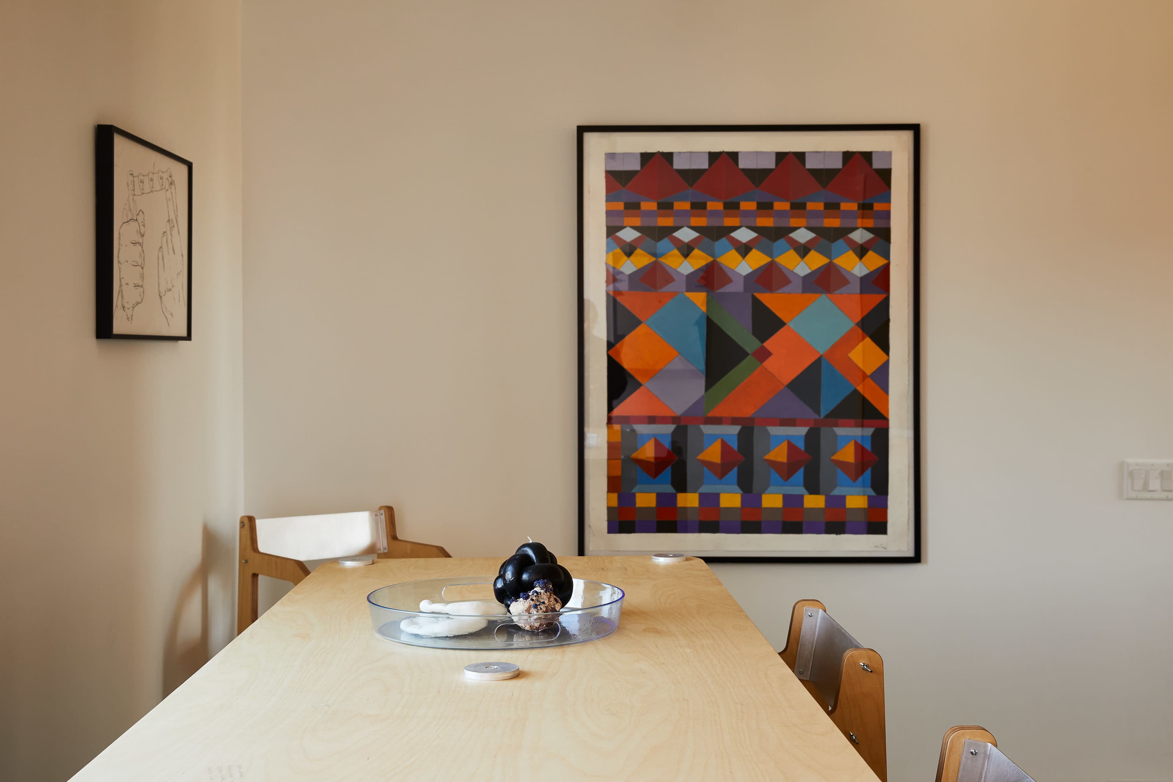 A dining table with a clear bowl of fruit is positioned in front of a colorful geometric artwork on the wall.