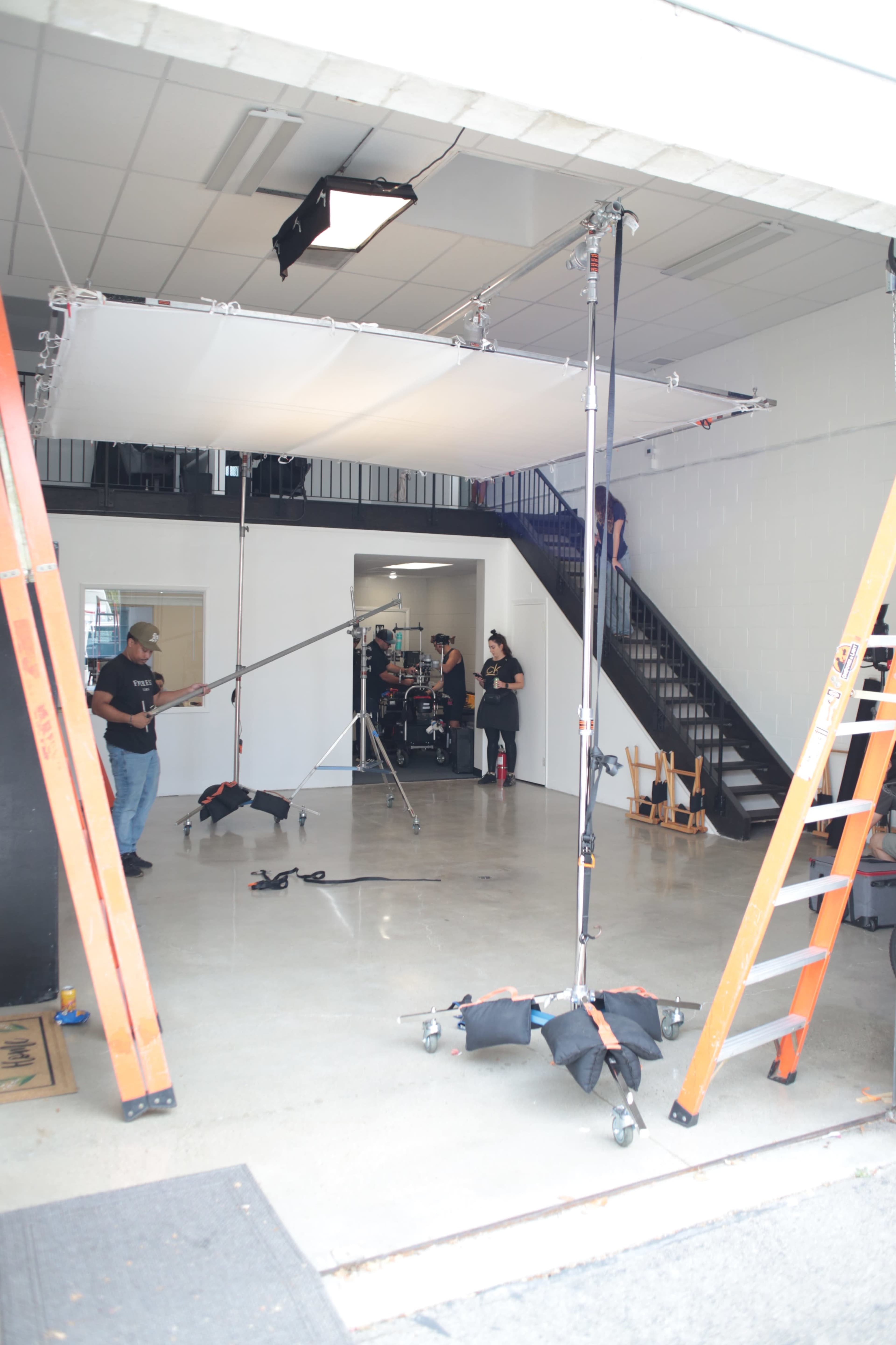 A film crew sets up lighting and equipment in a garage-like space with a staircase in the background.