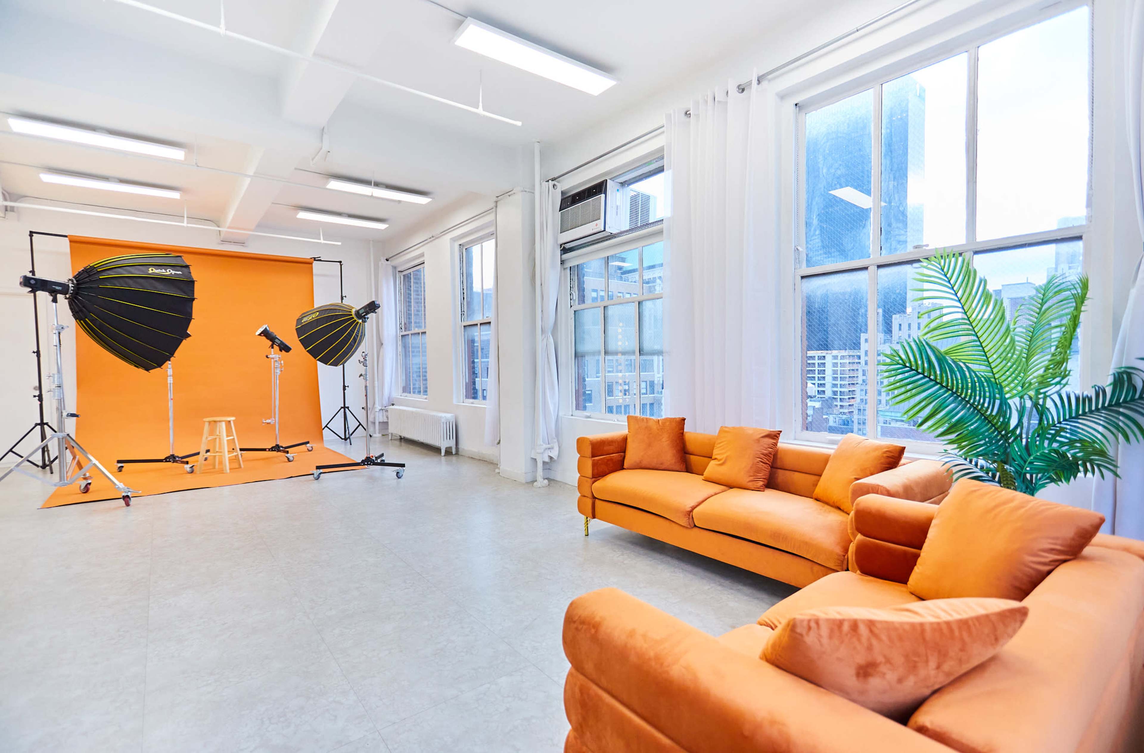 The image shows a brightly lit photography studio with a vibrant orange backdrop, two lighting setups, and an orange sofa in a corner near large windows.