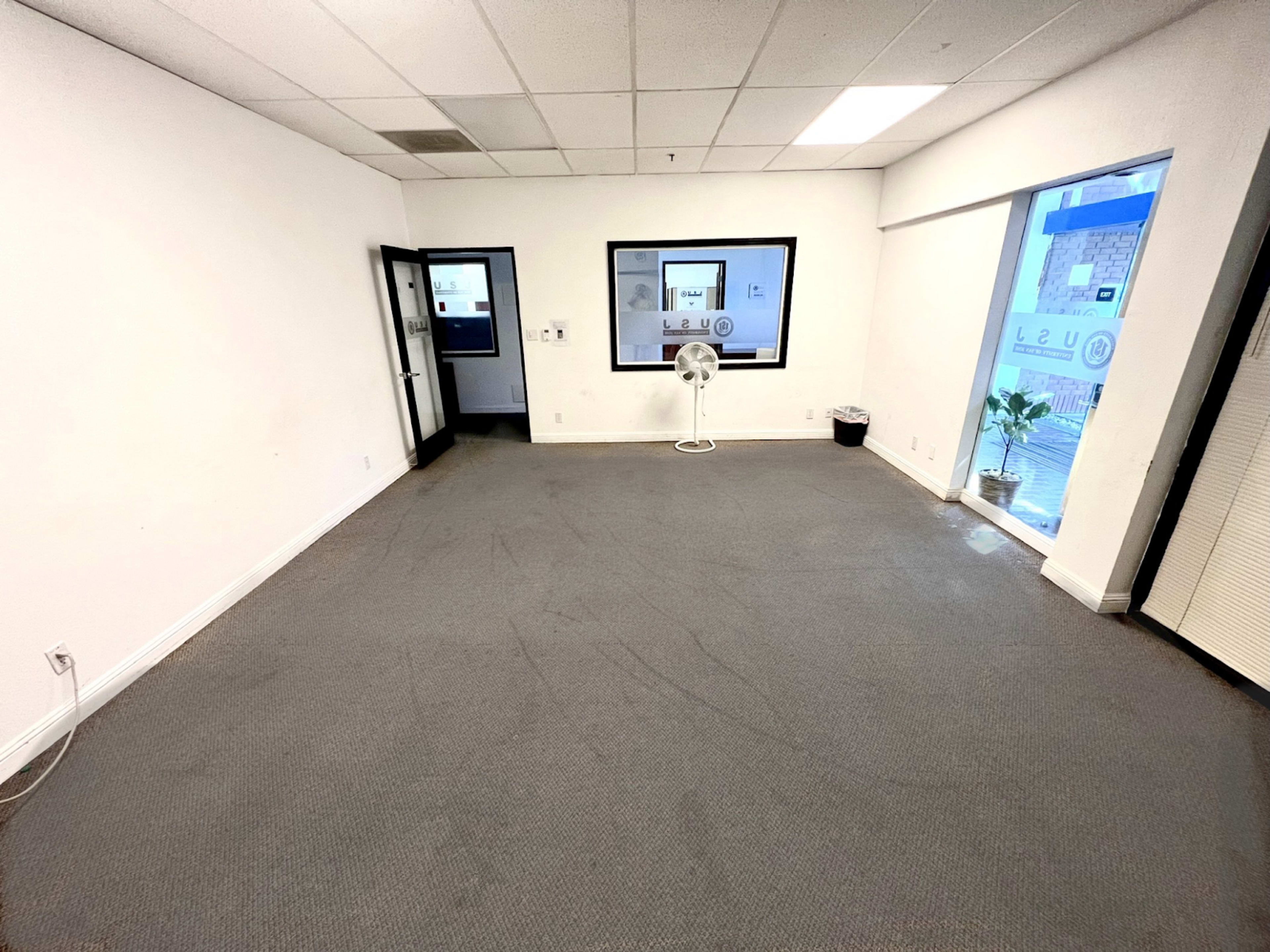 A bare, empty office space with gray carpeting and large windows allowing natural light to enter.