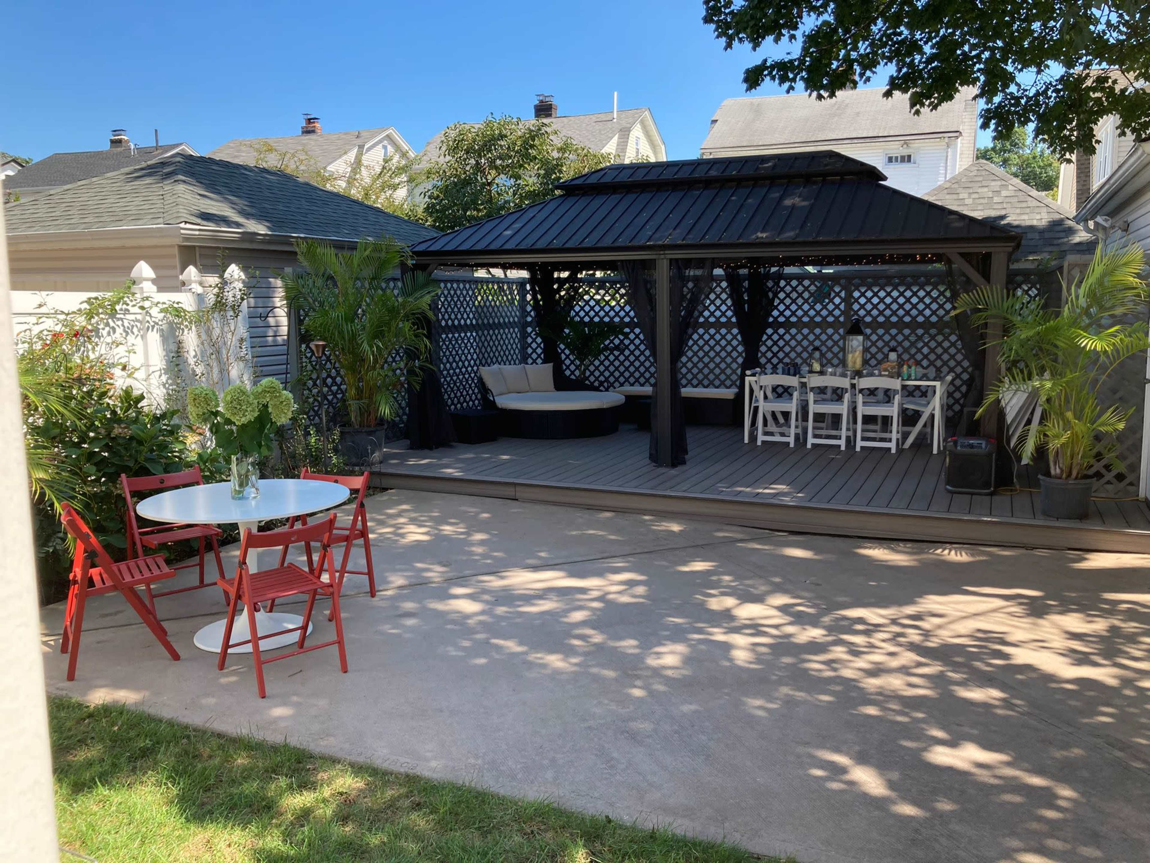 The image shows a landscaped backyard with a pergola, dining area, red chairs, and a hammock amidst surrounding greenery.