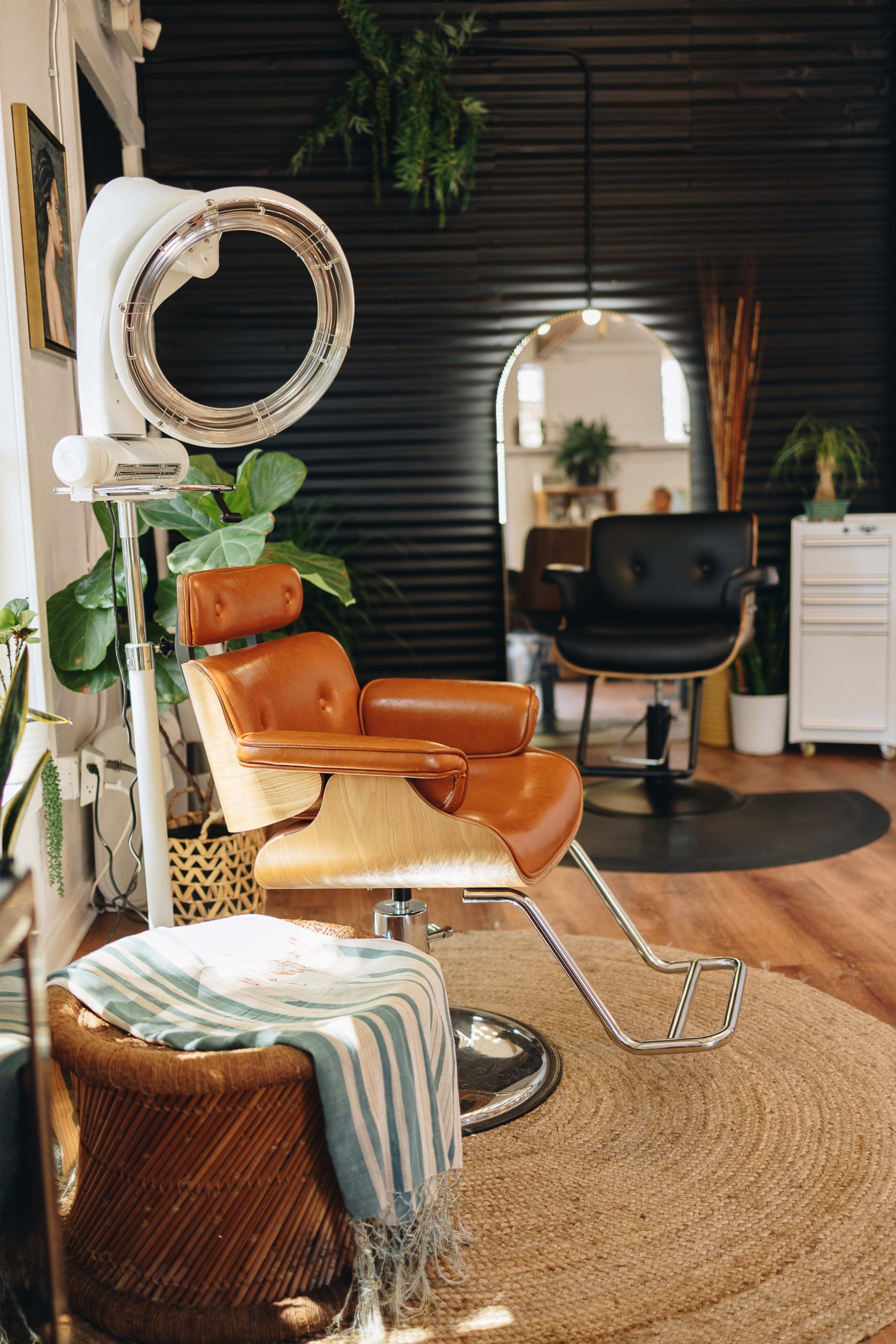 A modern hair salon interior featuring a brown leather styling chair, a round mirror, and lush green plants.