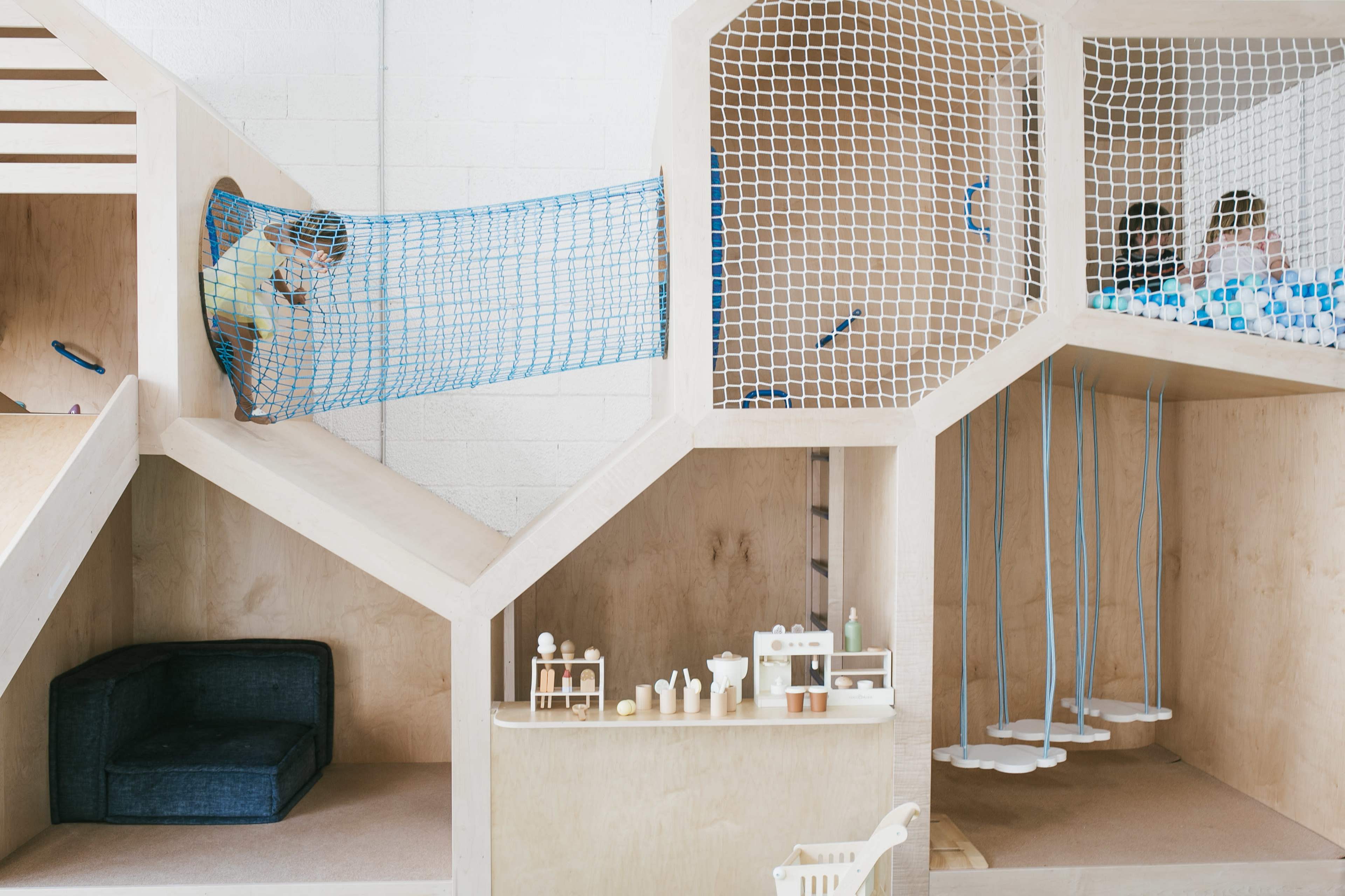 The image shows a children's play area featuring a wooden structure with climbing nets, seating, and various play elements designed for interactive activities.