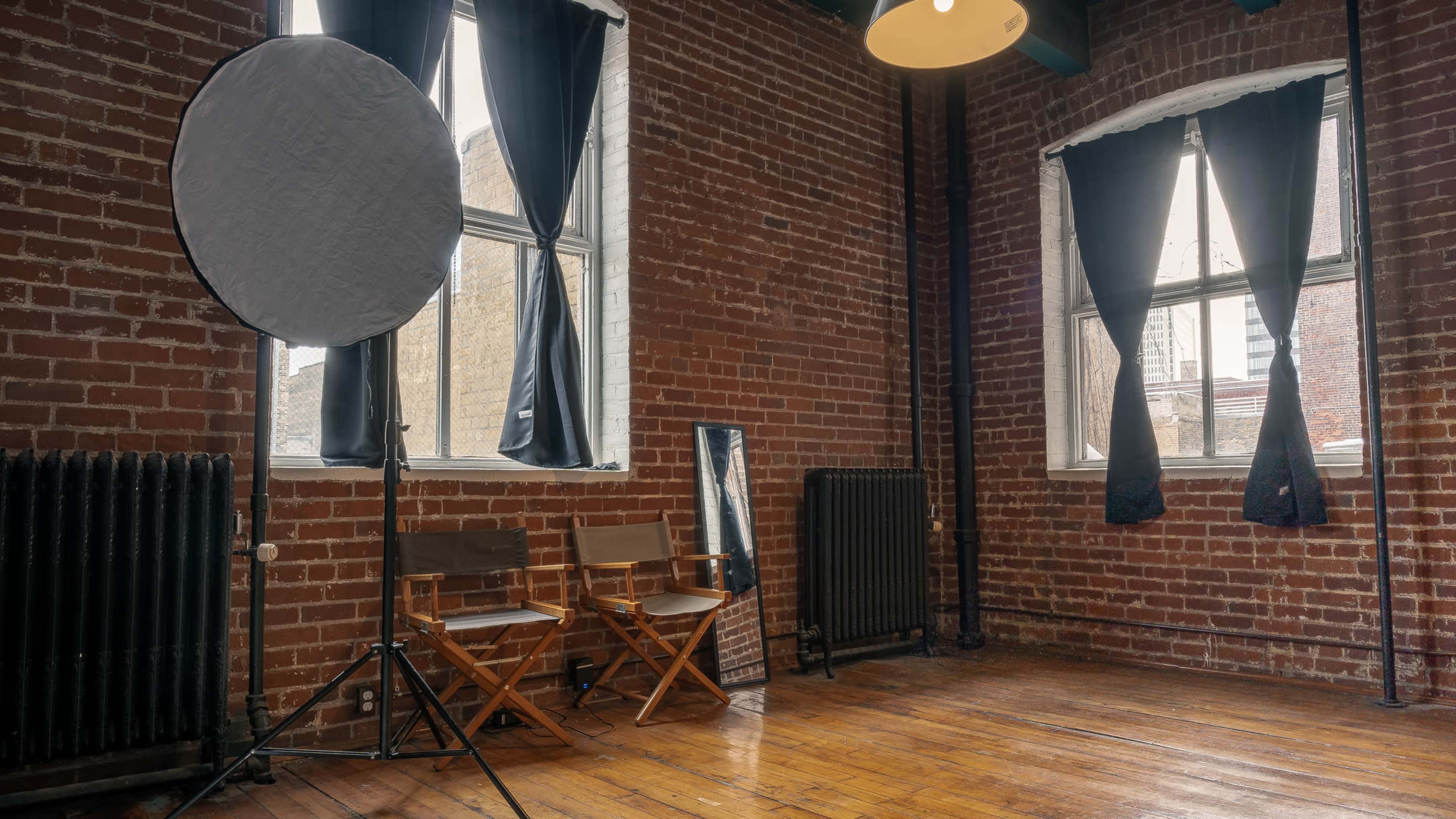 An empty room with brick walls, two director's chairs, a large reflector, and black curtains over the windows.