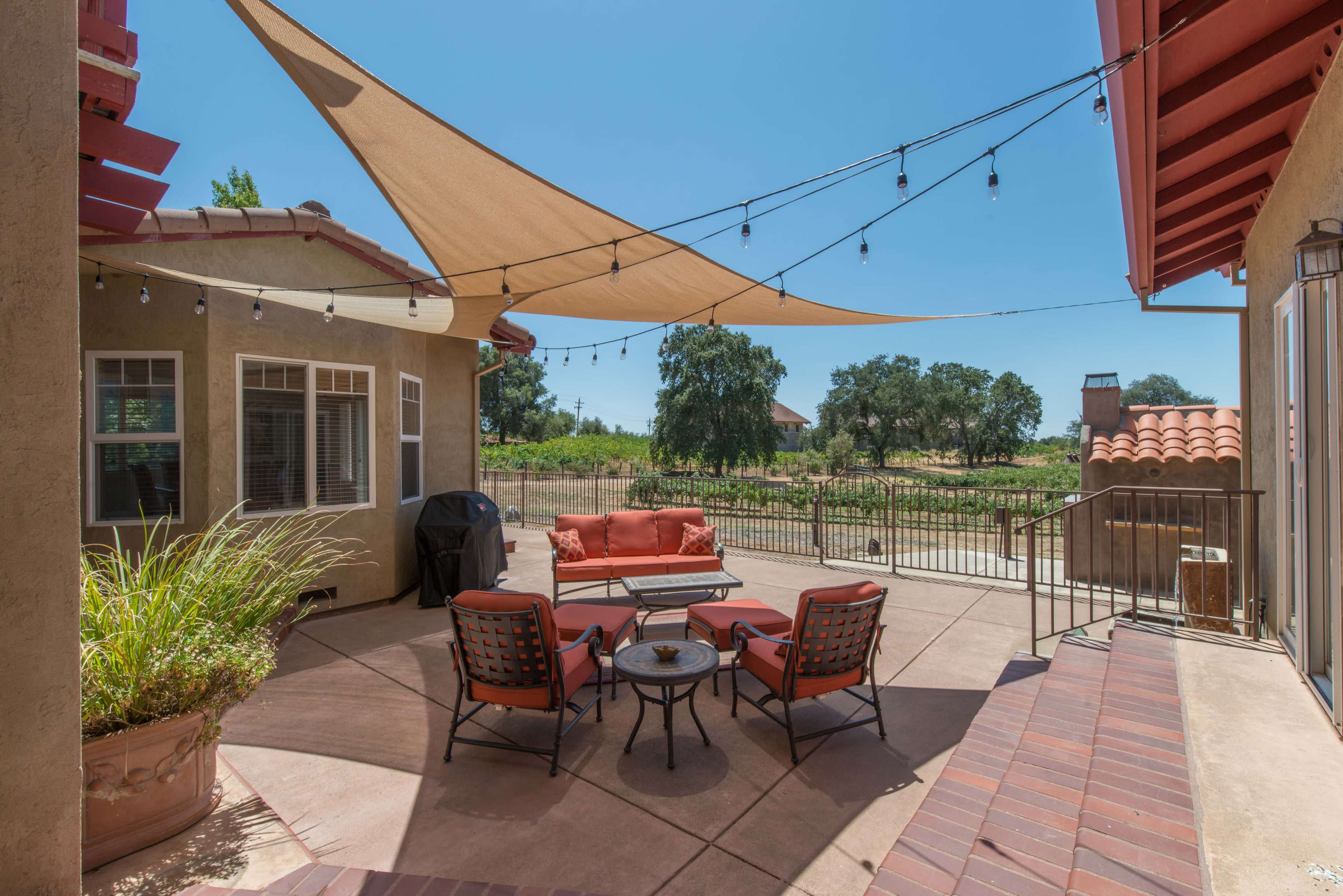 A patio area with a seating arrangement, shaded by a sail, overlooks a vineyard.