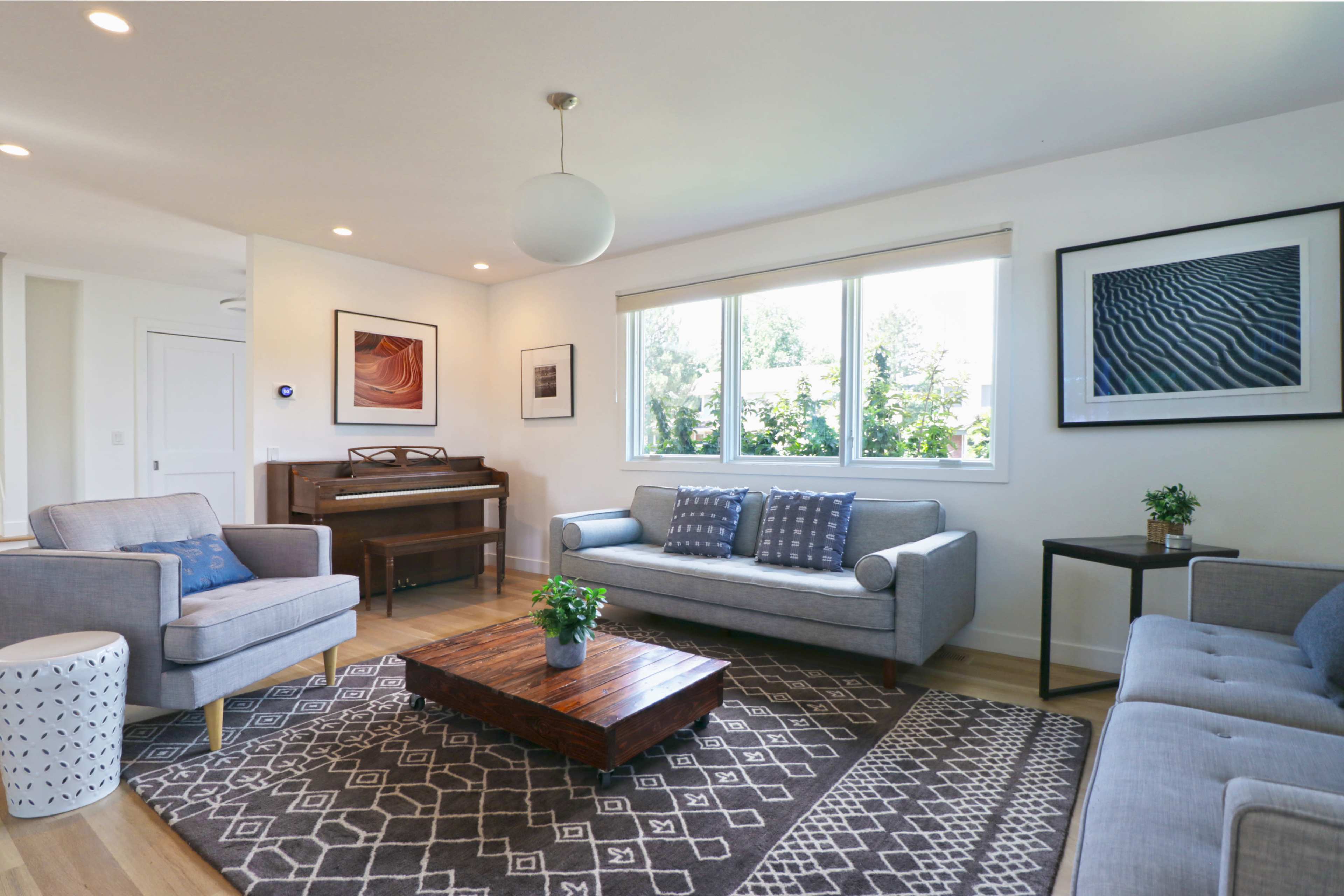 A bright and modern living room features a piano, two gray sofas, a wooden coffee table, and large windows offering a view of greenery.