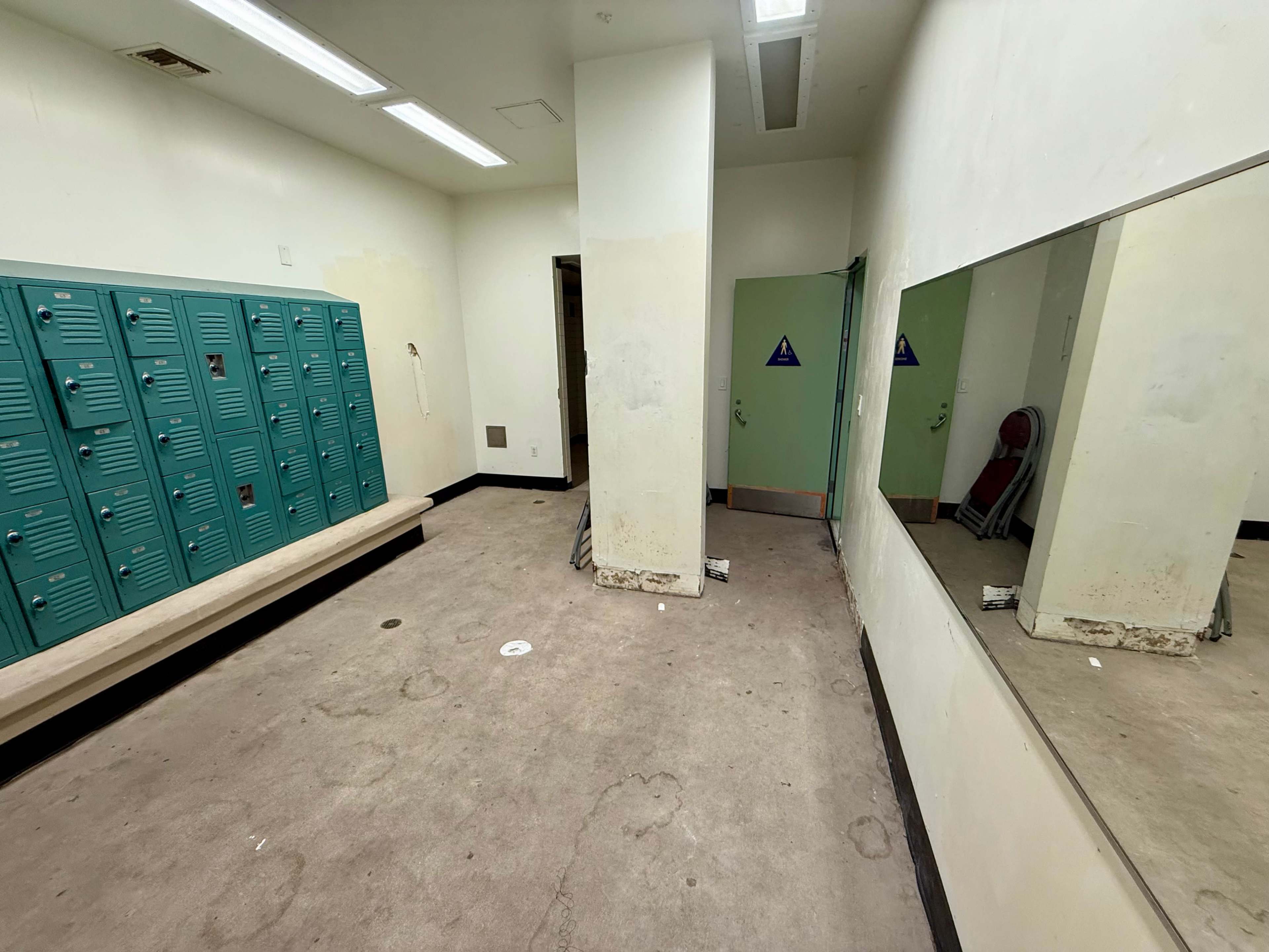 The image shows a sparsely furnished locker room with teal lockers lining one wall, a bench, and two mirrors reflecting the empty space.