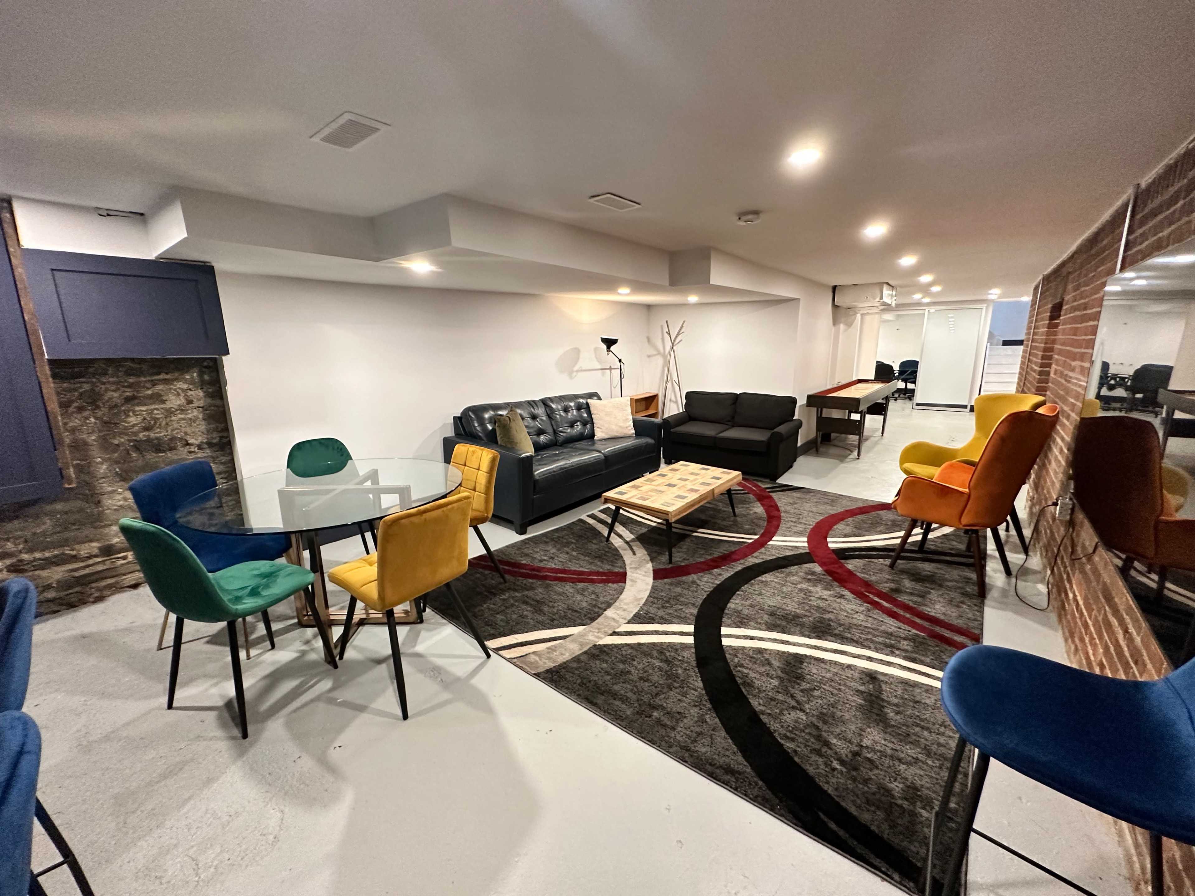 A modern, semi-open basement lounge area featuring a mix of seating options, including colorful chairs and a glass dining table, alongside a pool table and a decorative rug.