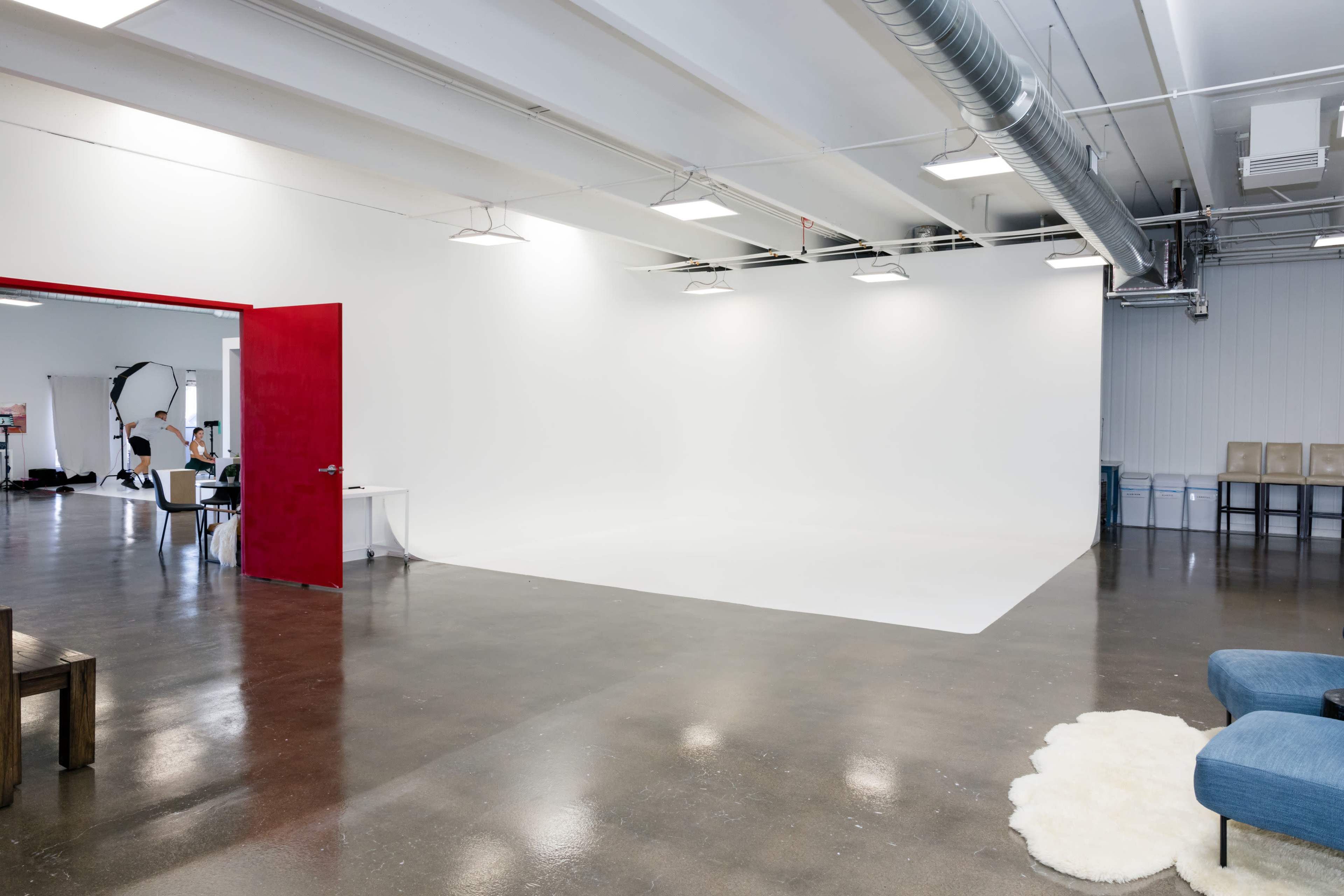 The image shows a spacious, minimalist studio with a bright white backdrop and a red door leading to another area.