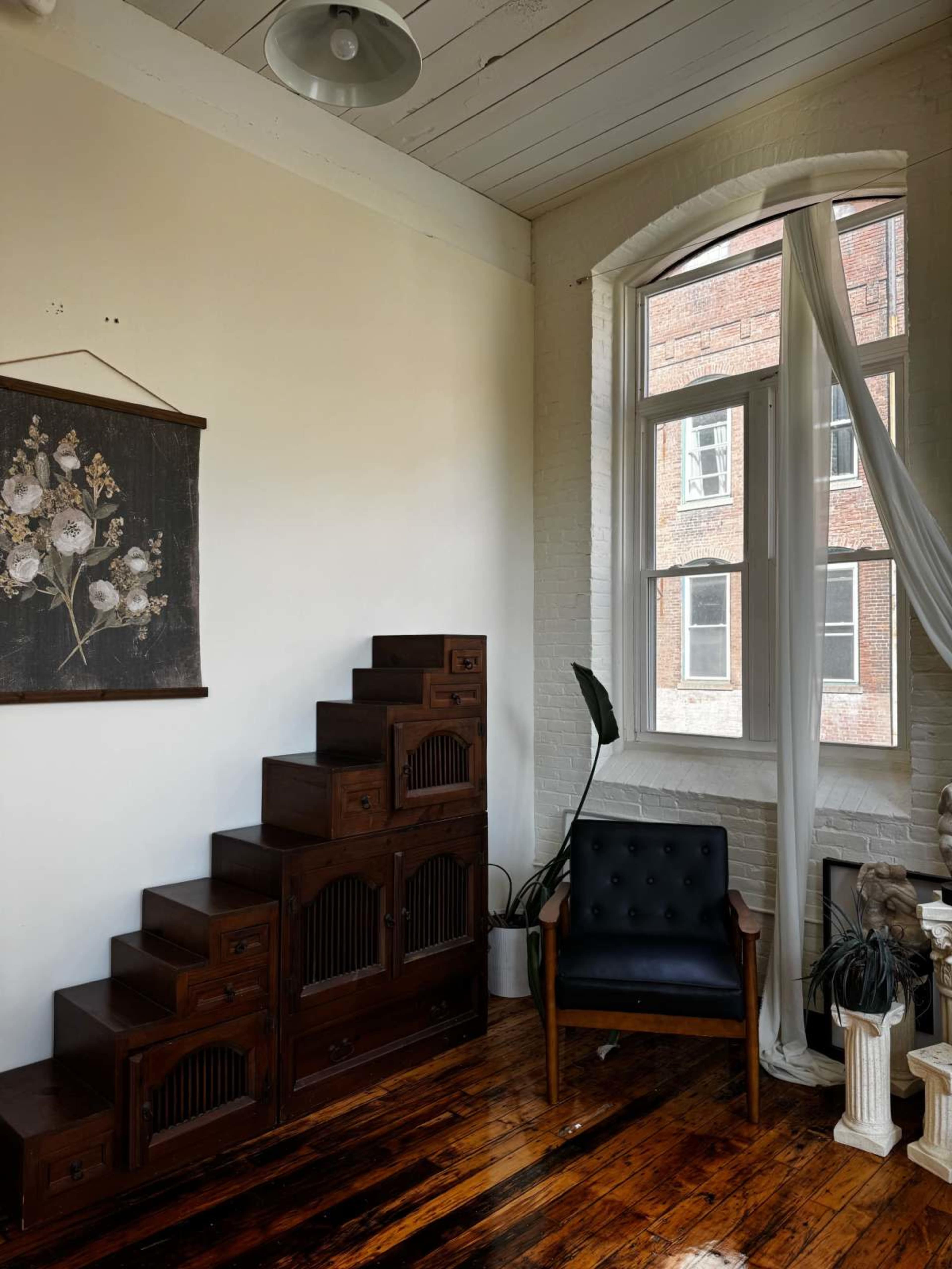 The room features a wooden staircase cabinet, a black leather chair, a window with sheer curtains, and a floral wall hanging.