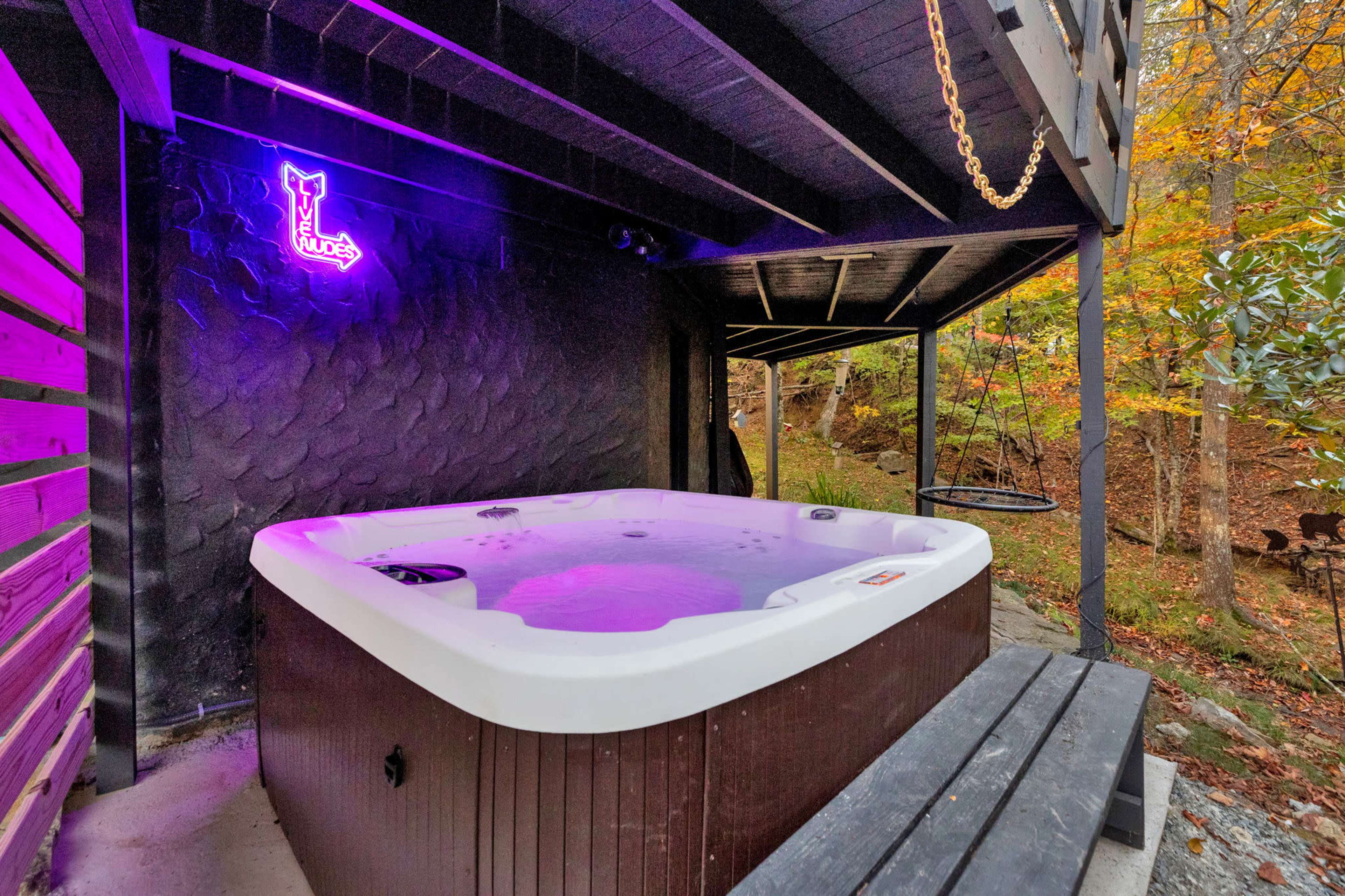 A hot tub with purple lighting is situated under a covered wooden structure, surrounded by trees and a rocky path.