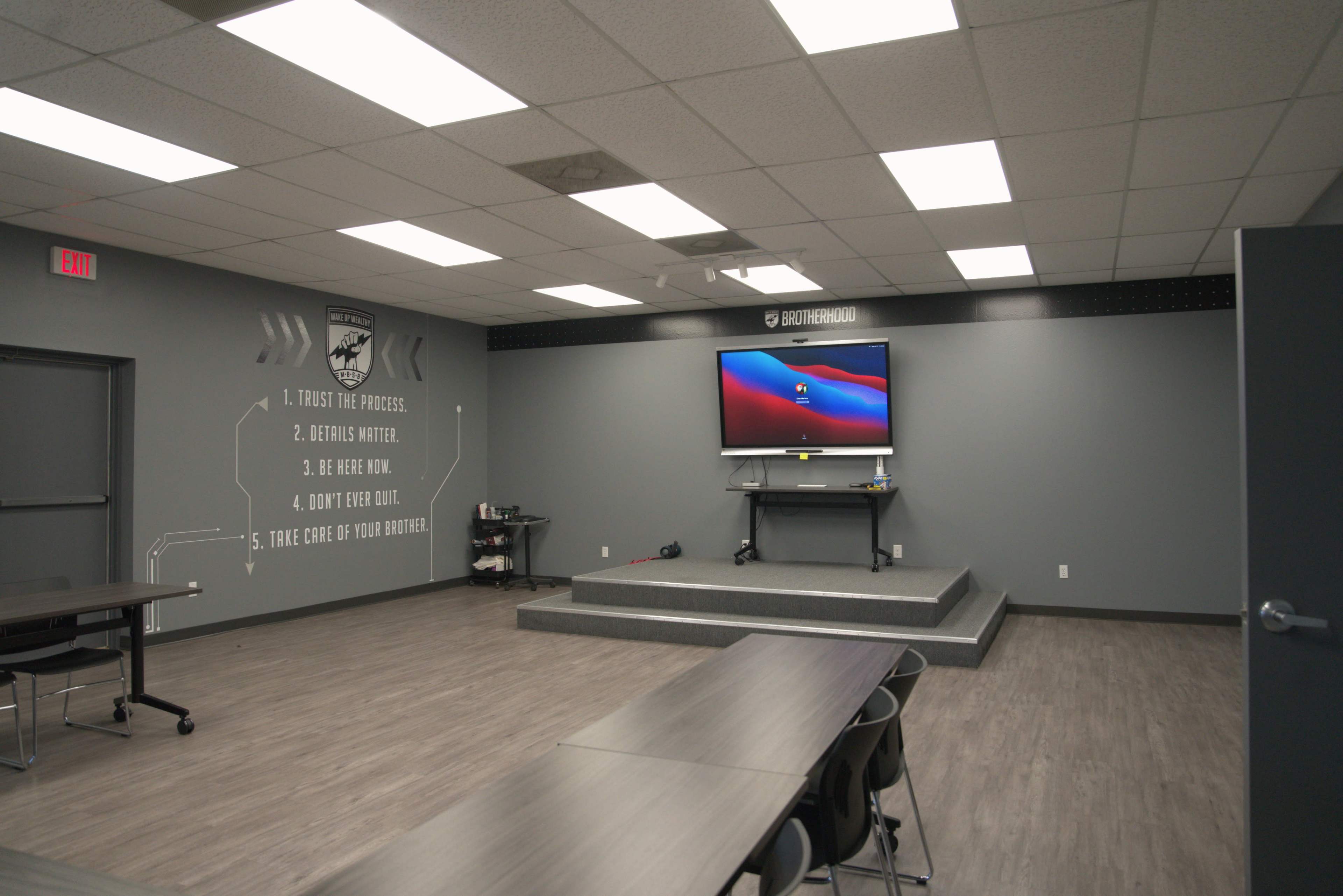 The image shows a minimalist classroom or meeting room with a stage for presentations, a mounted screen, and several tables arranged in rows.