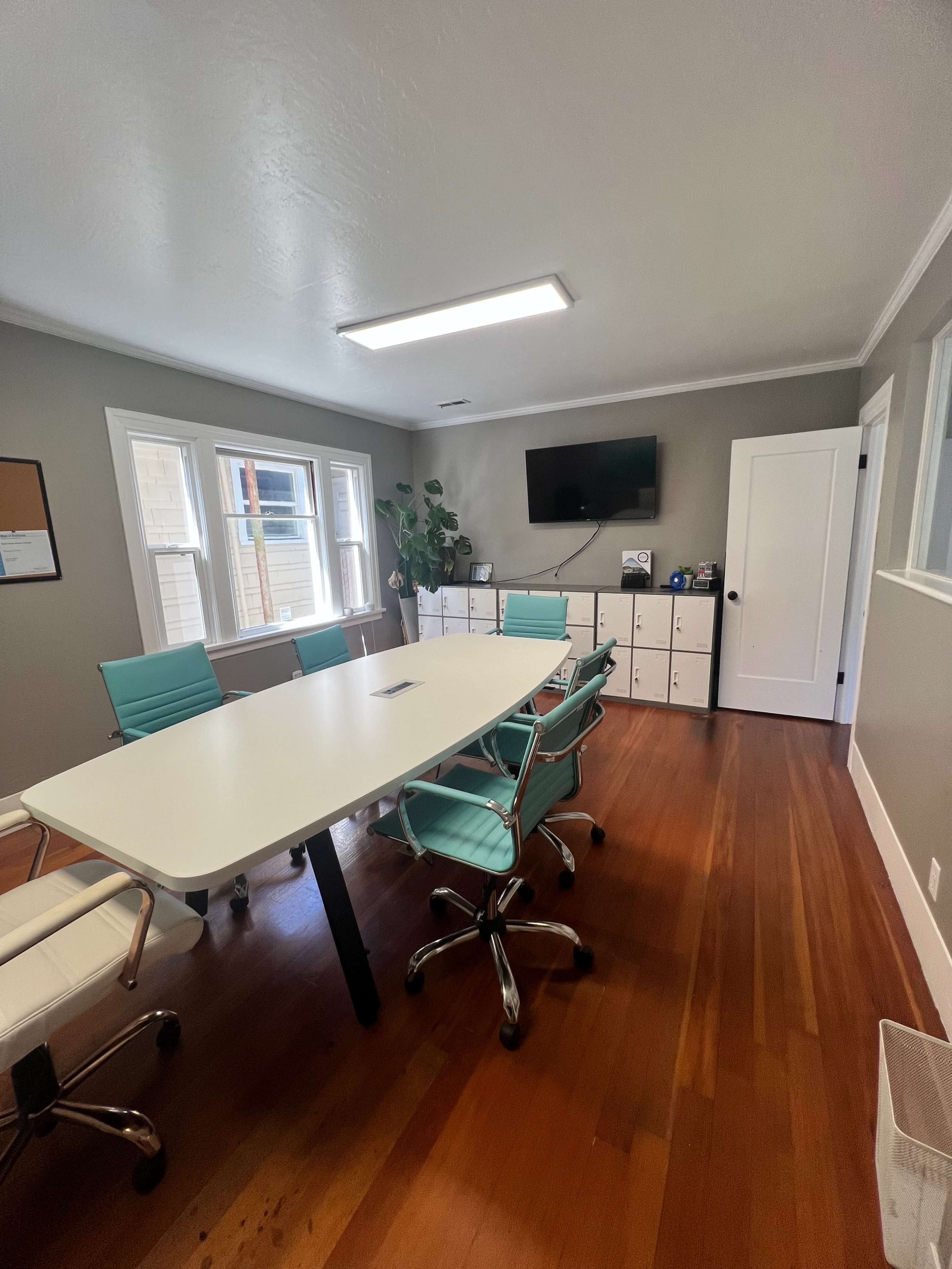 The image shows a modern conference room with a large white table, turquoise chairs, and a television mounted on the wall, accompanied by storage cabinets.