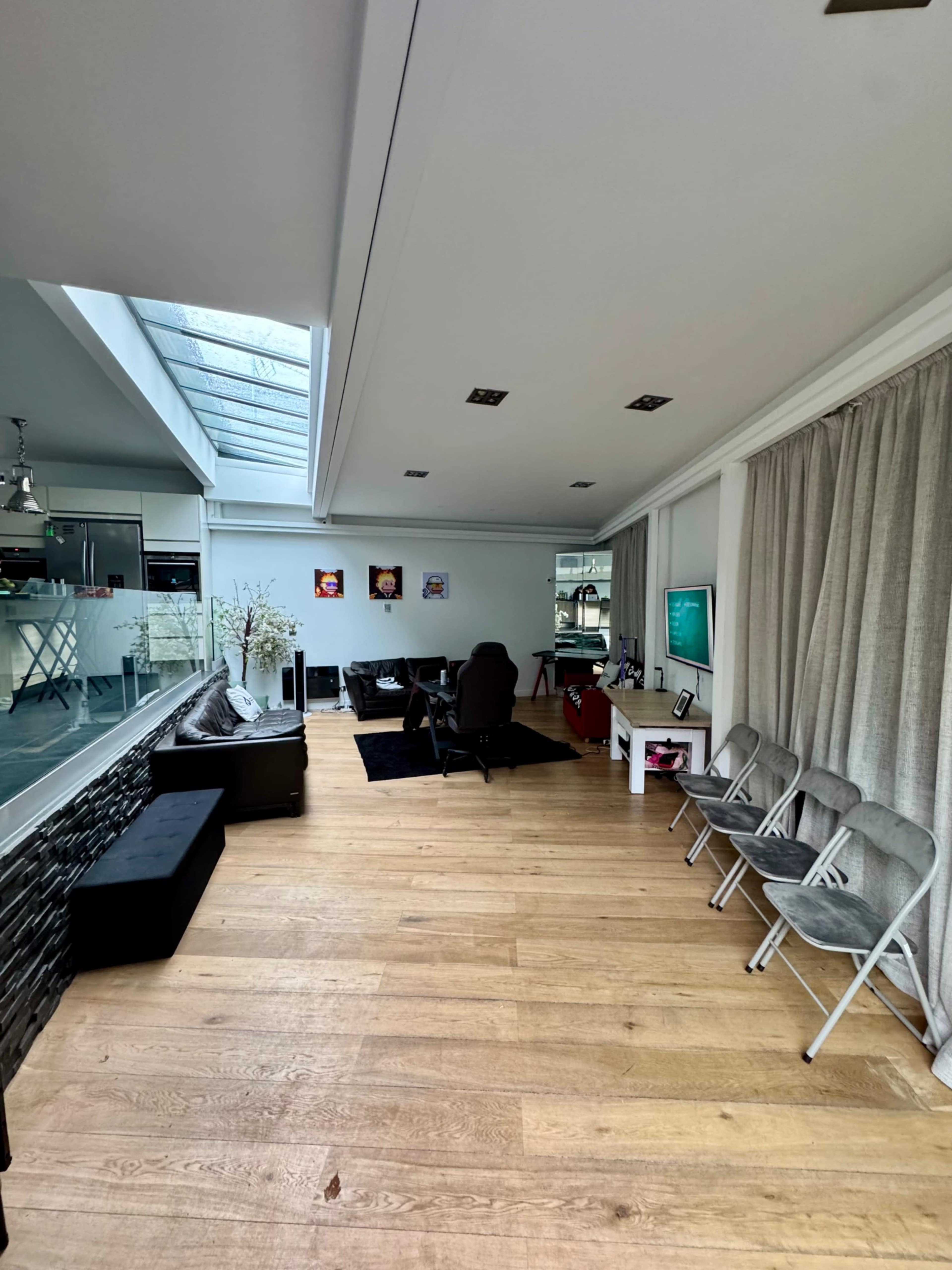 The image shows a modern, spacious living area with a glass ceiling, wooden flooring, a desk with a chair, and several folding chairs arranged along one side.