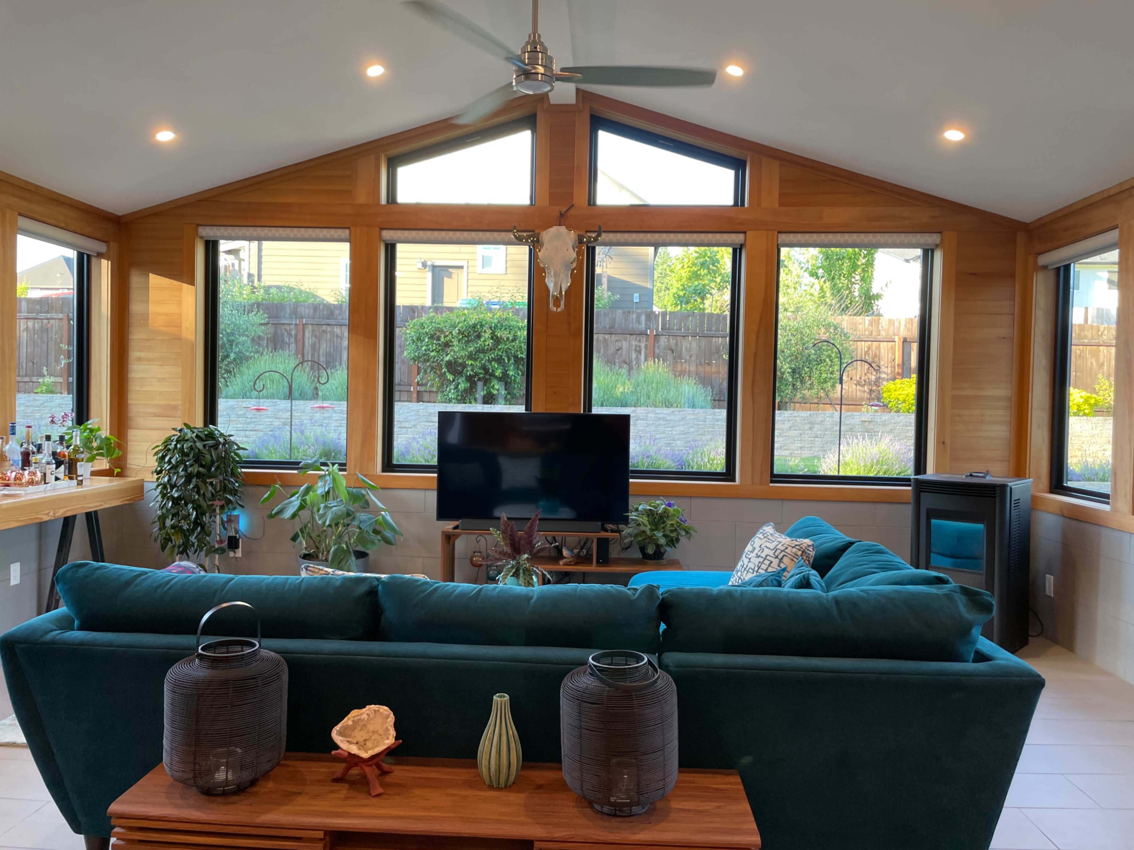 A modern sunroom with large windows, a television, and a green sofa, surrounded by various indoor plants and decorative items on a wooden table.