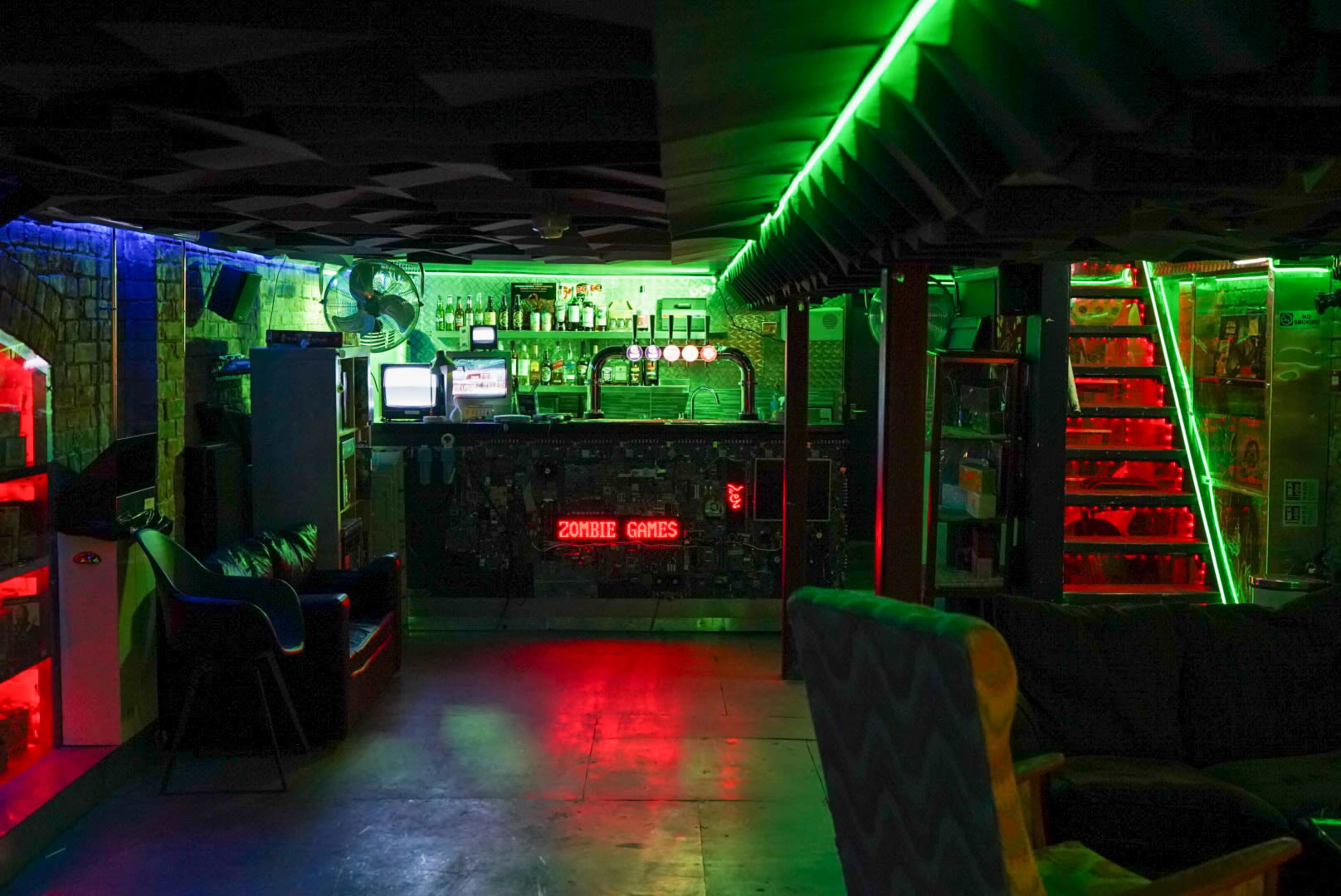 The image shows a dimly lit game-themed lounge with neon green and red lighting, featuring a bar, shelves of games, and stairs leading to an upper level.