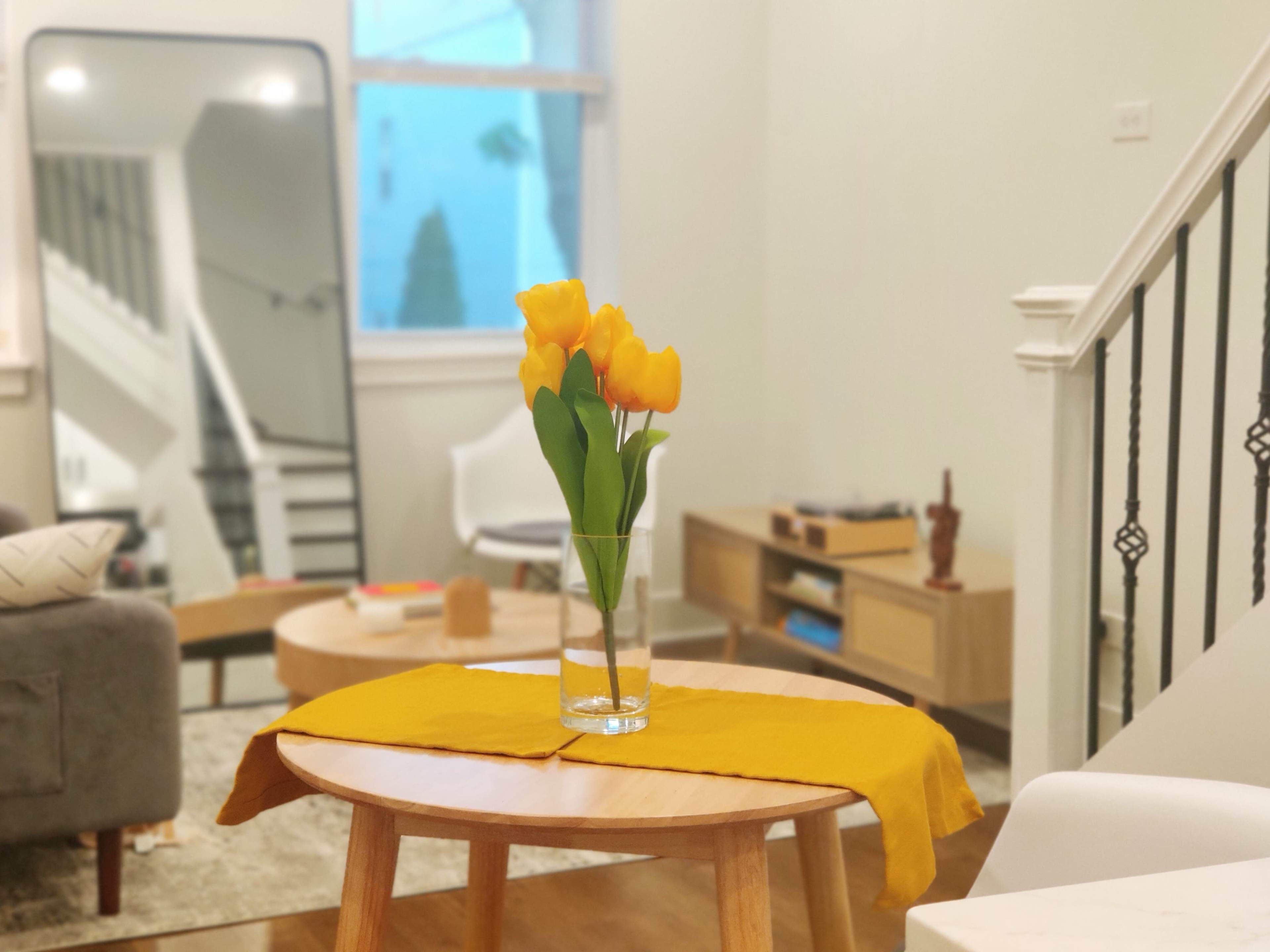 A vase of yellow tulips sits on a round wooden coffee table, accented by a yellow table runner, in a living room with a mirror and light-colored furniture.