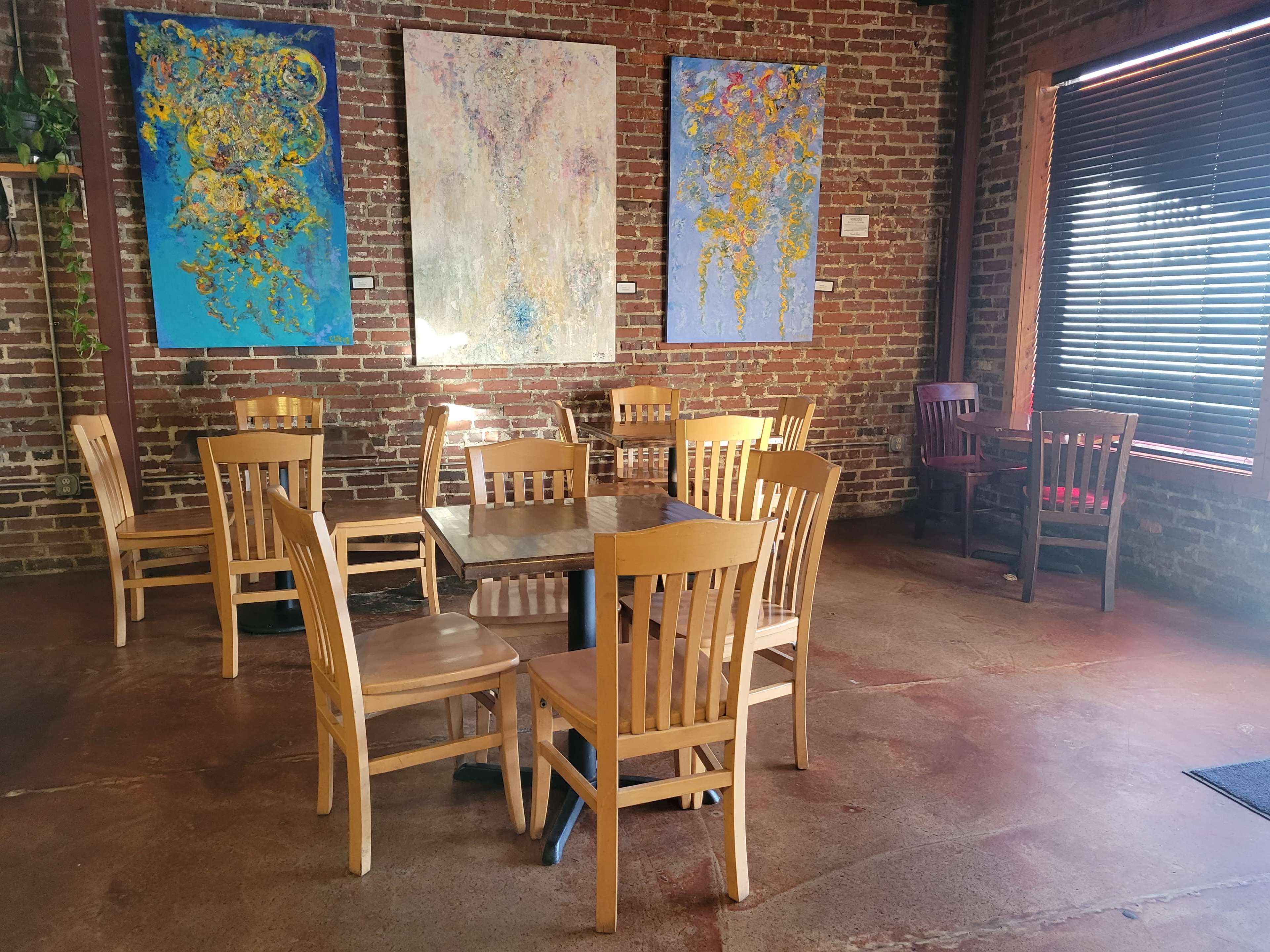 A cafe interior with wooden tables and chairs arranged on a polished floor, accompanied by colorful abstract paintings on the brick walls.