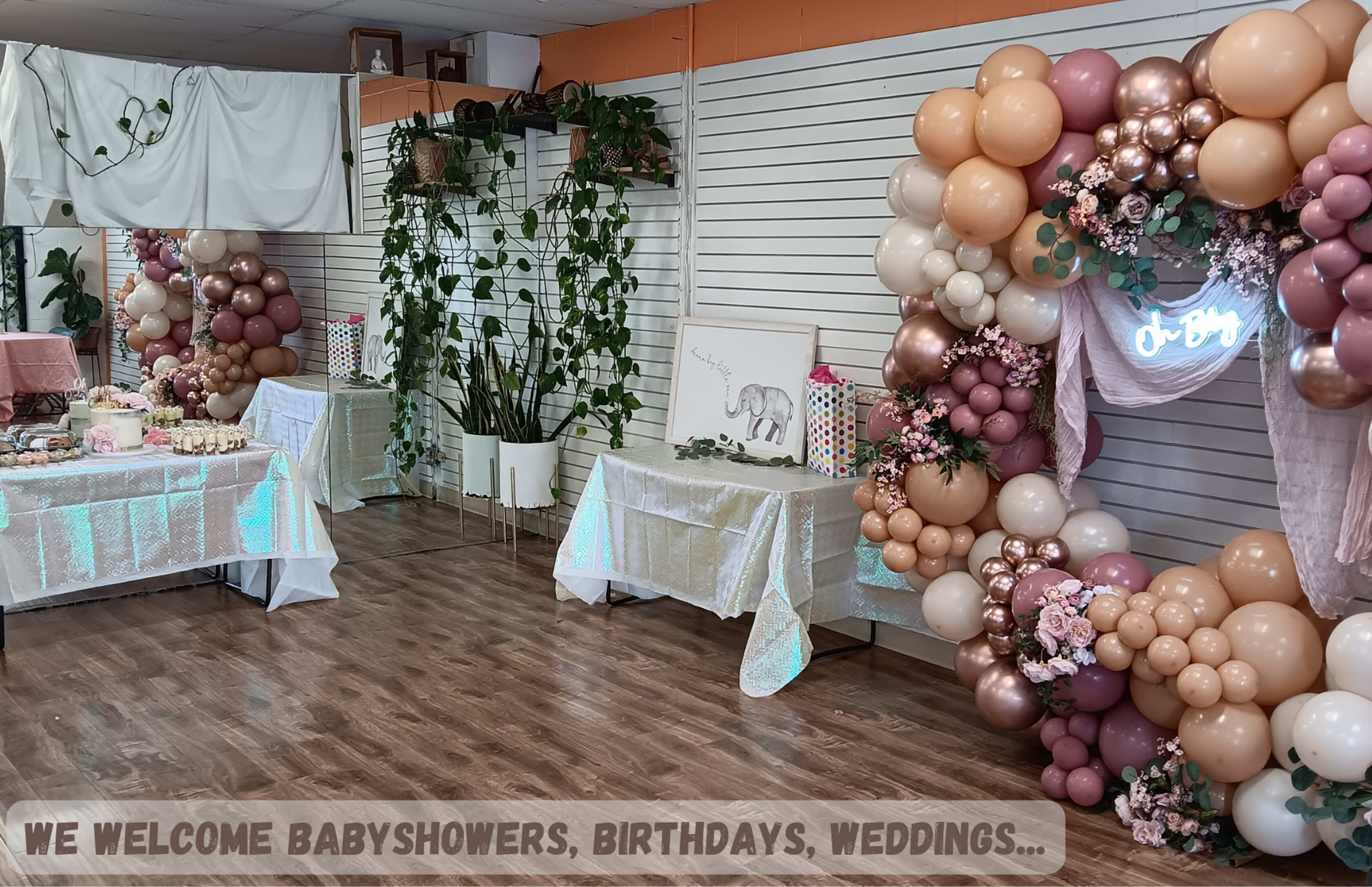 A decorated indoor space featuring tables with desserts and a balloon arch adorned with flowers, designed for events like baby showers and birthdays.
