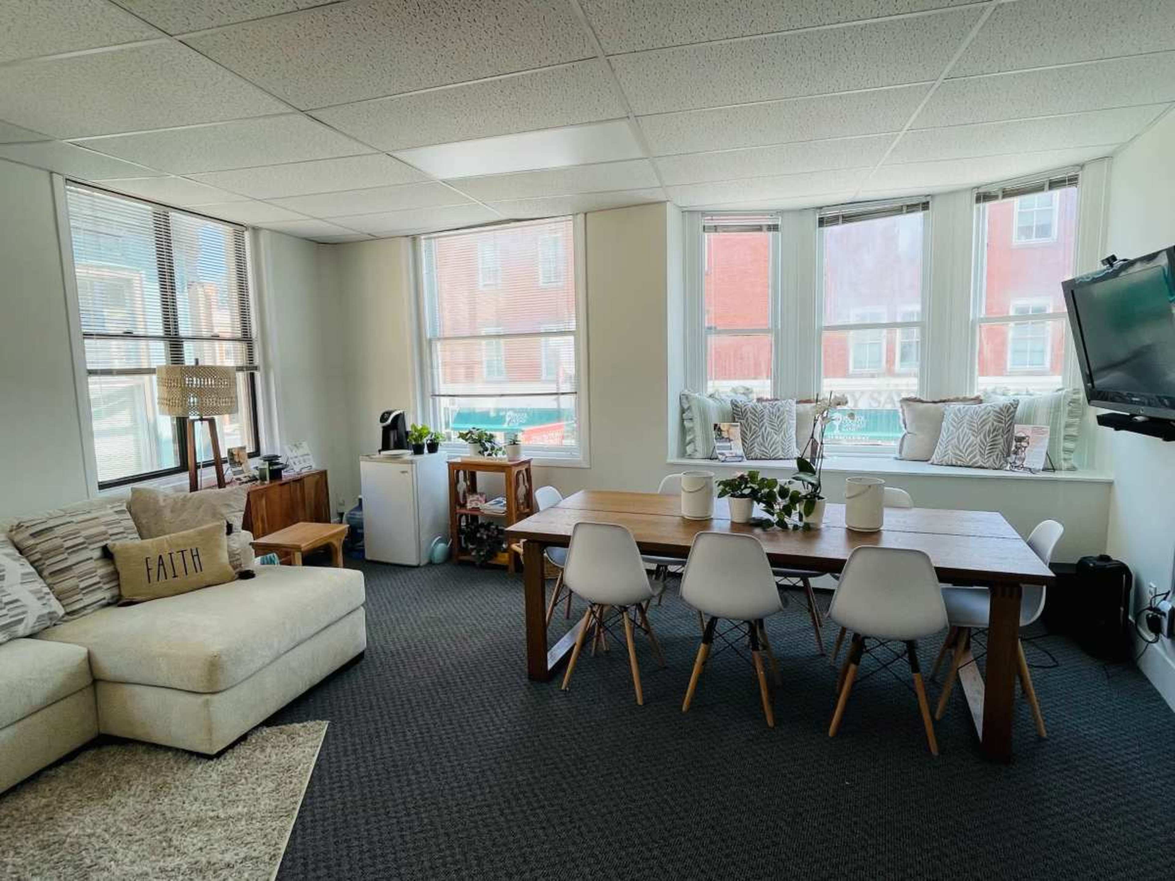 The room features a seating area with a light-colored couch, a wooden dining table surrounded by white chairs, and large windows allowing natural light.