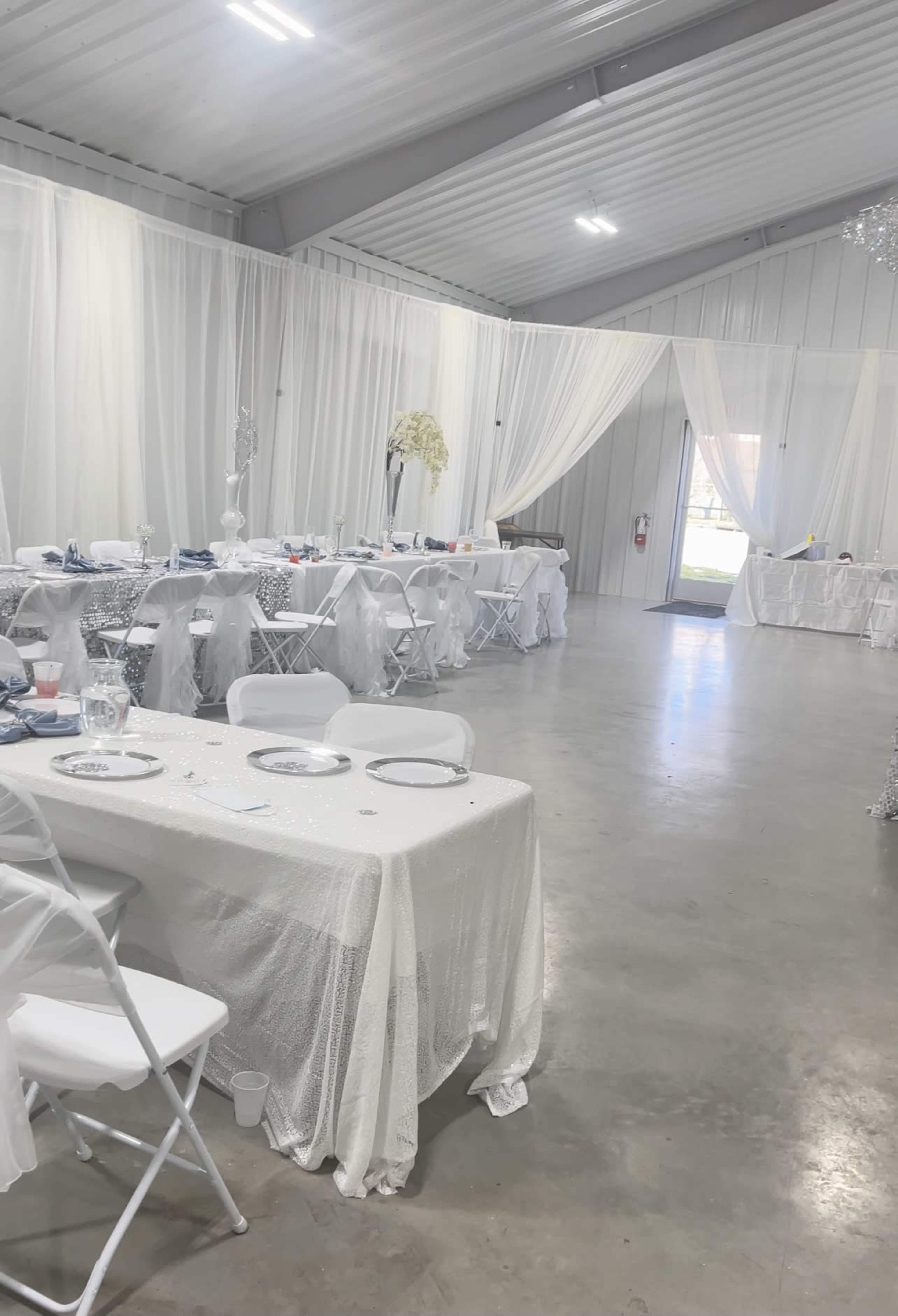 A large indoor event space is set up with white tables and chairs, adorned with decorative tablecloths and surrounded by sheer white drapery.