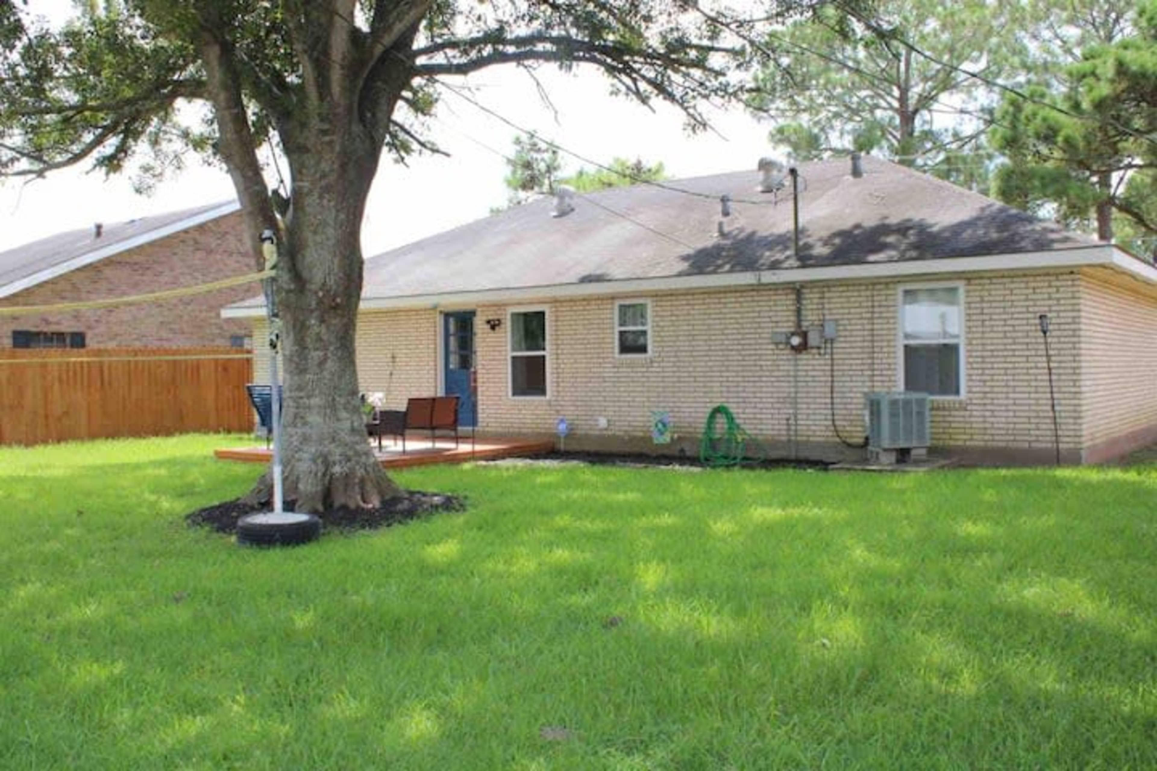 The image shows a single-story house with a tan brick exterior situated in a grassy yard, featuring a patio area with outdoor furniture and an air conditioning unit.