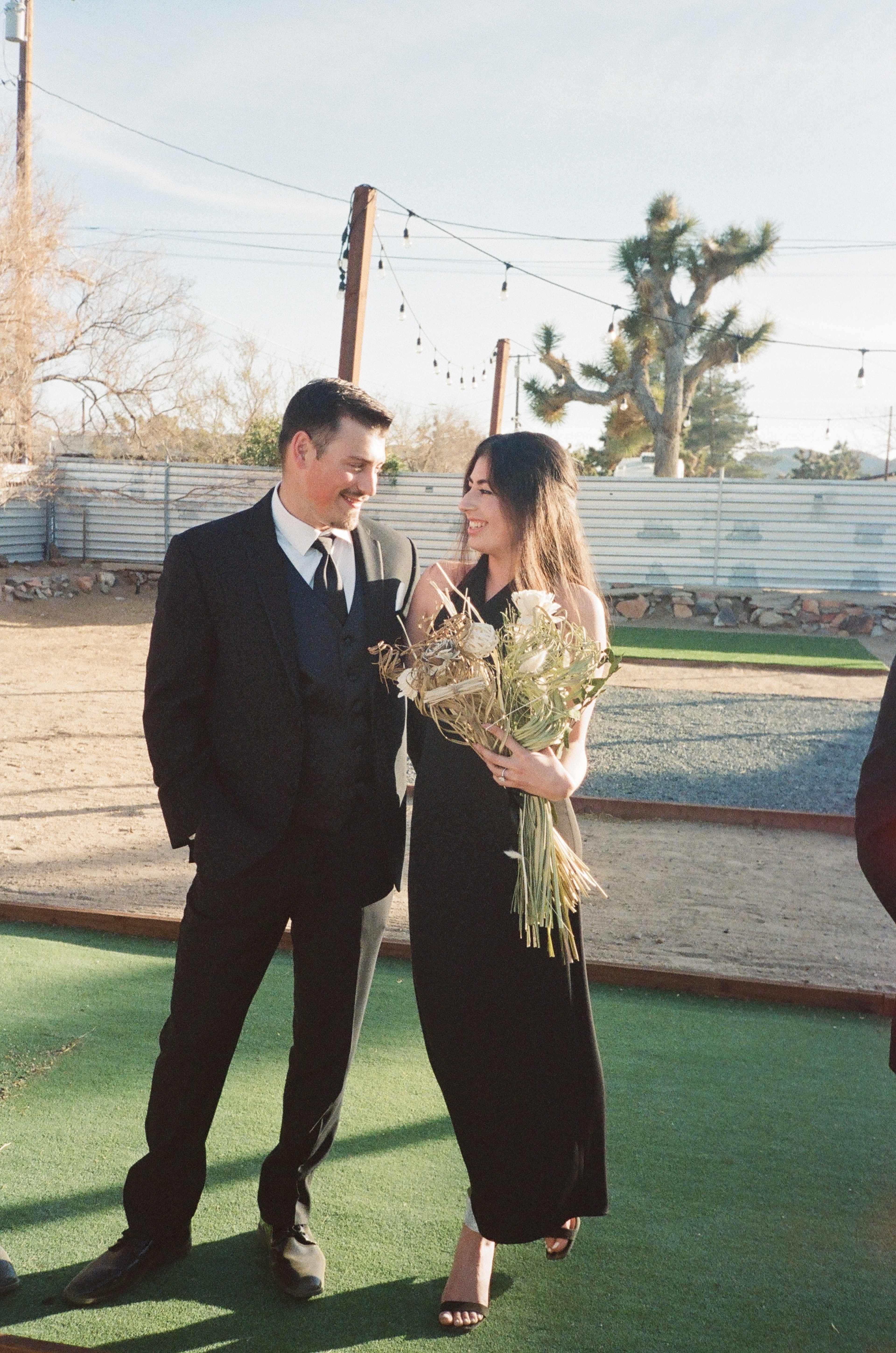 A man in a suit walks alongside a woman in a black dress, both smiling, while she holds a bouquet of dried flowers against a desert landscape.
