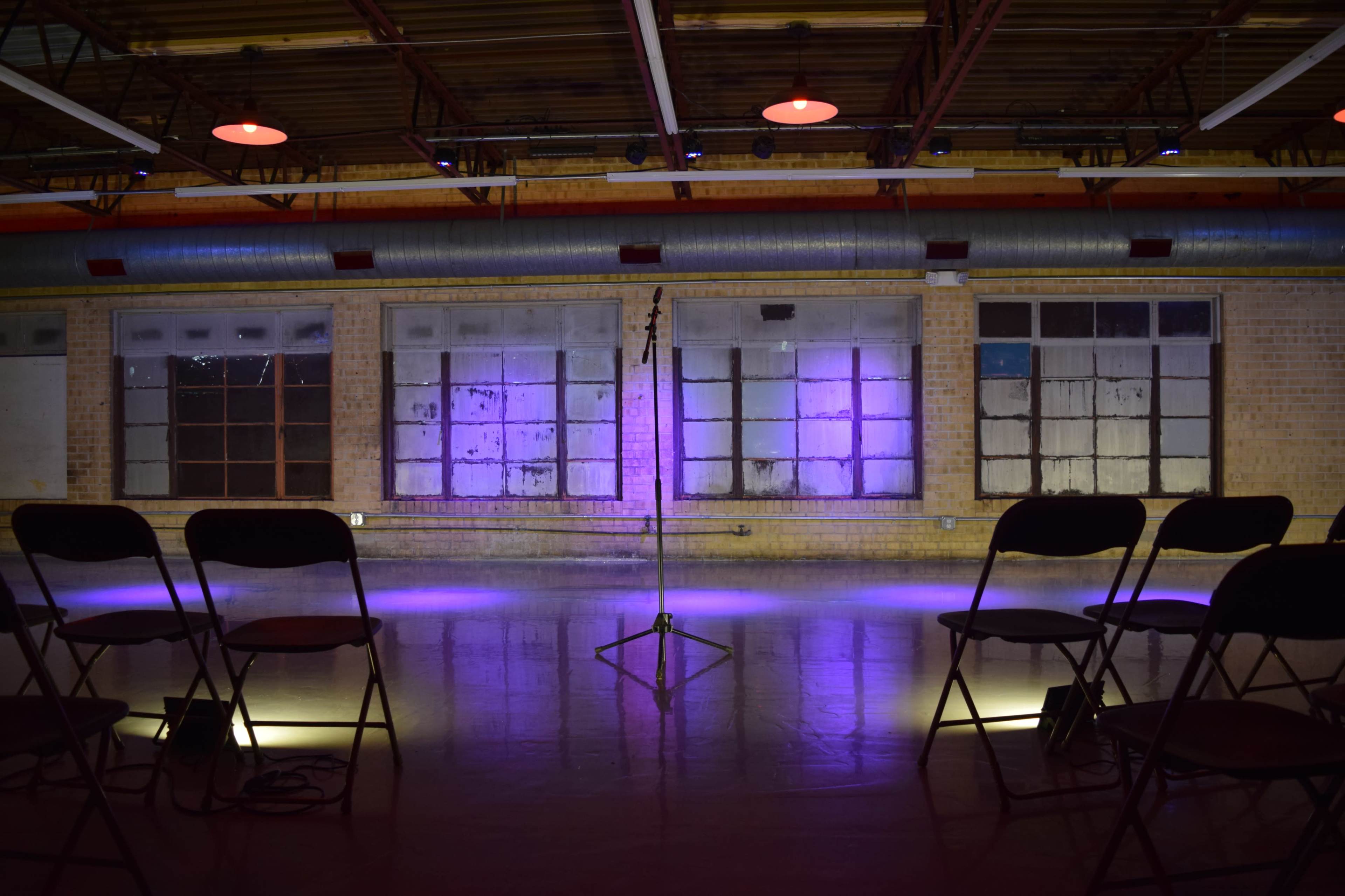 An empty performance space with a microphone stand at the center, surrounded by chairs and illuminated by purple lighting.