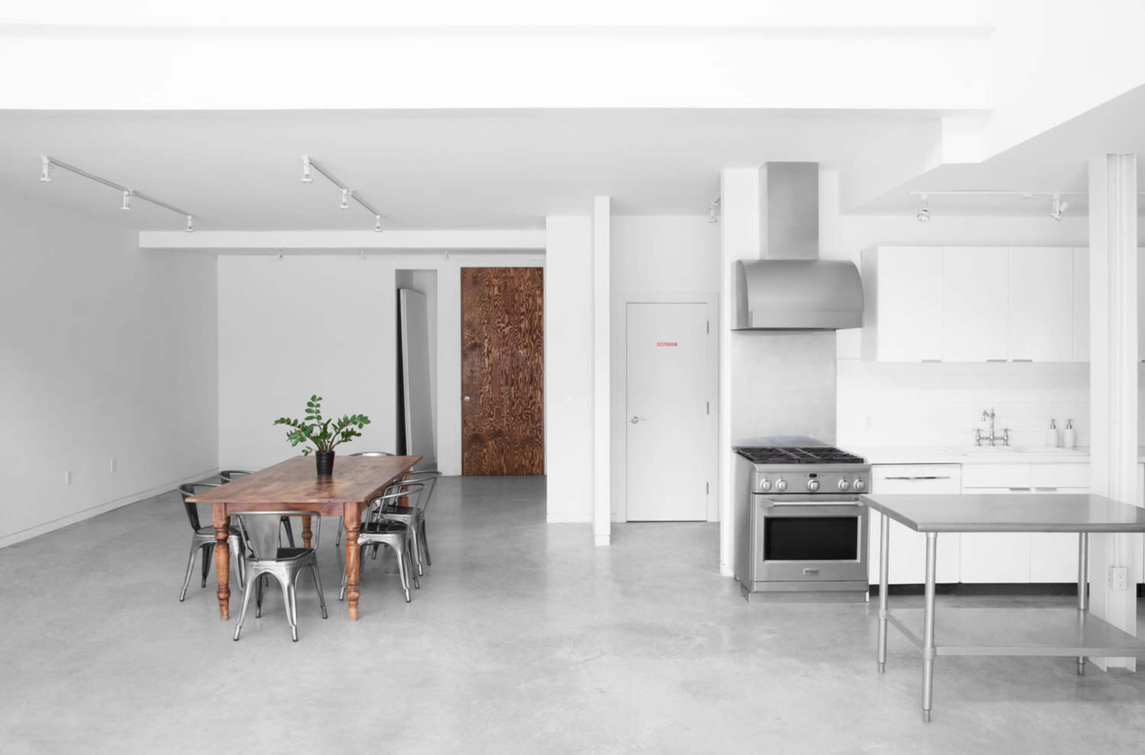A minimalist kitchen and dining area features a wooden table, metal chairs, and stainless steel appliances in a bright, open space with concrete flooring.