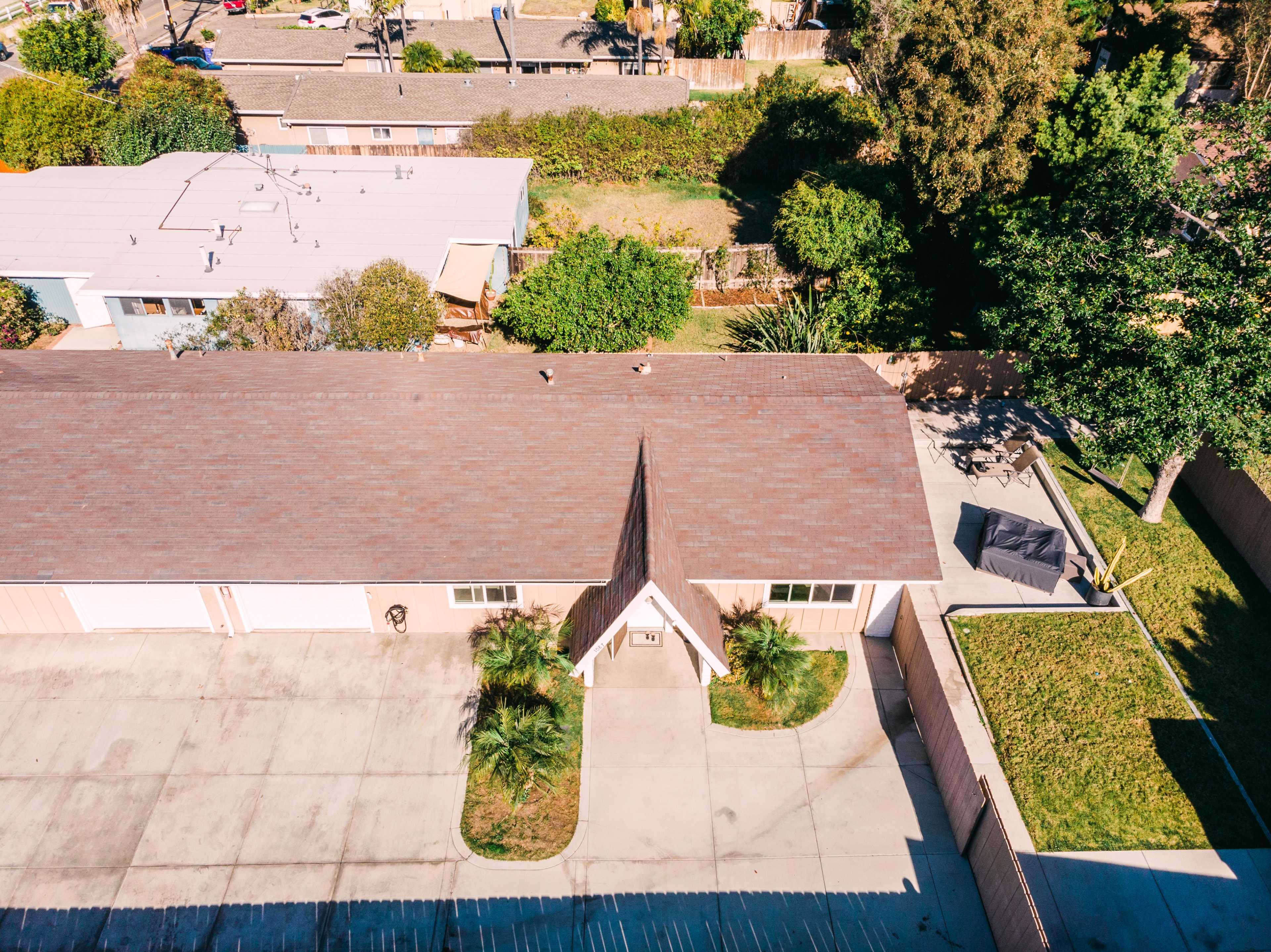 The image shows an aerial view of a residential property with a pitched roof, surrounded by greenery and adjacent structures in a suburban area.