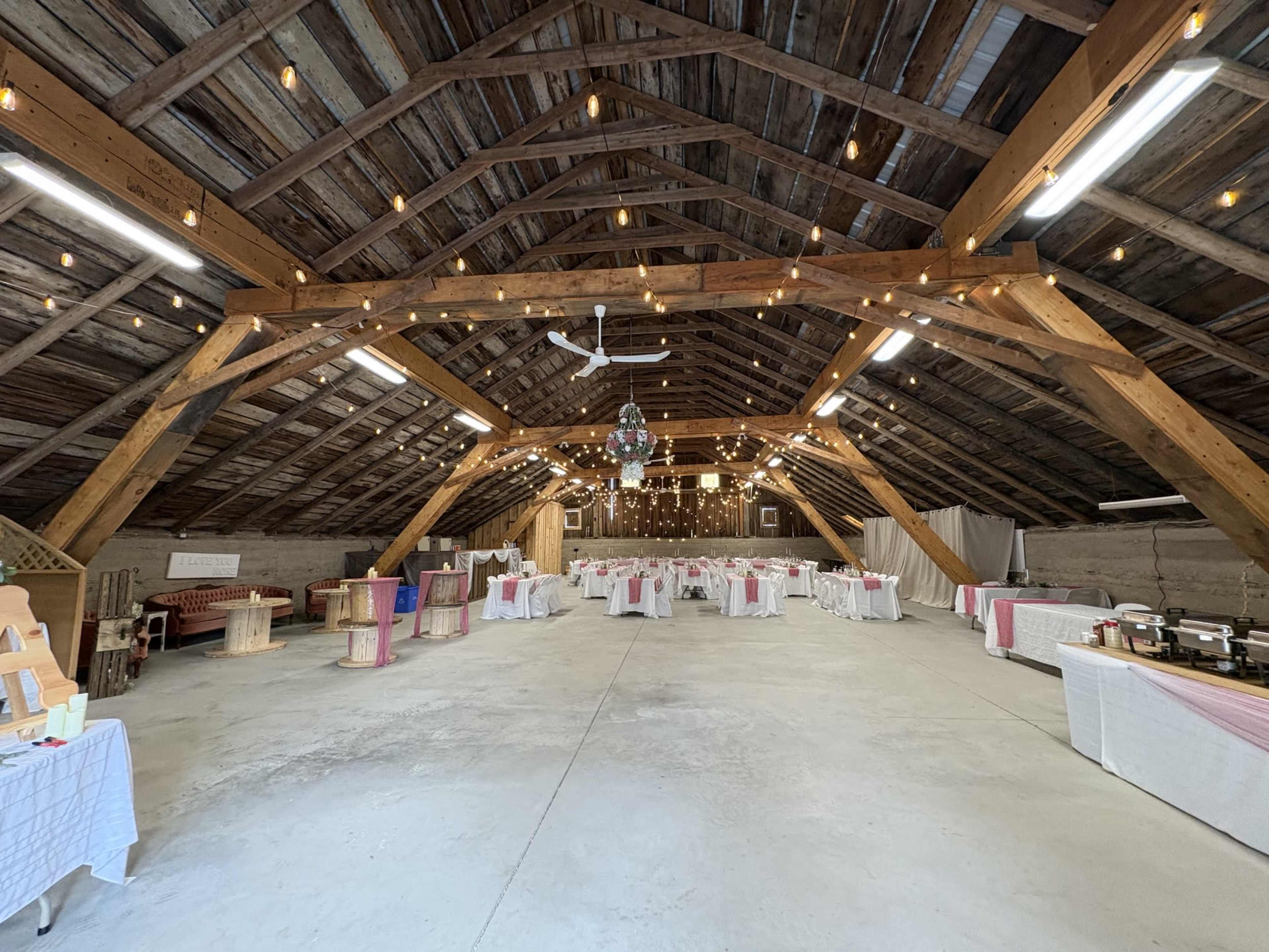 The image shows a spacious barn interior adorned with wooden beams, featuring tables set for an event with white tablecloths and pink accents, along with decorative lighting.
