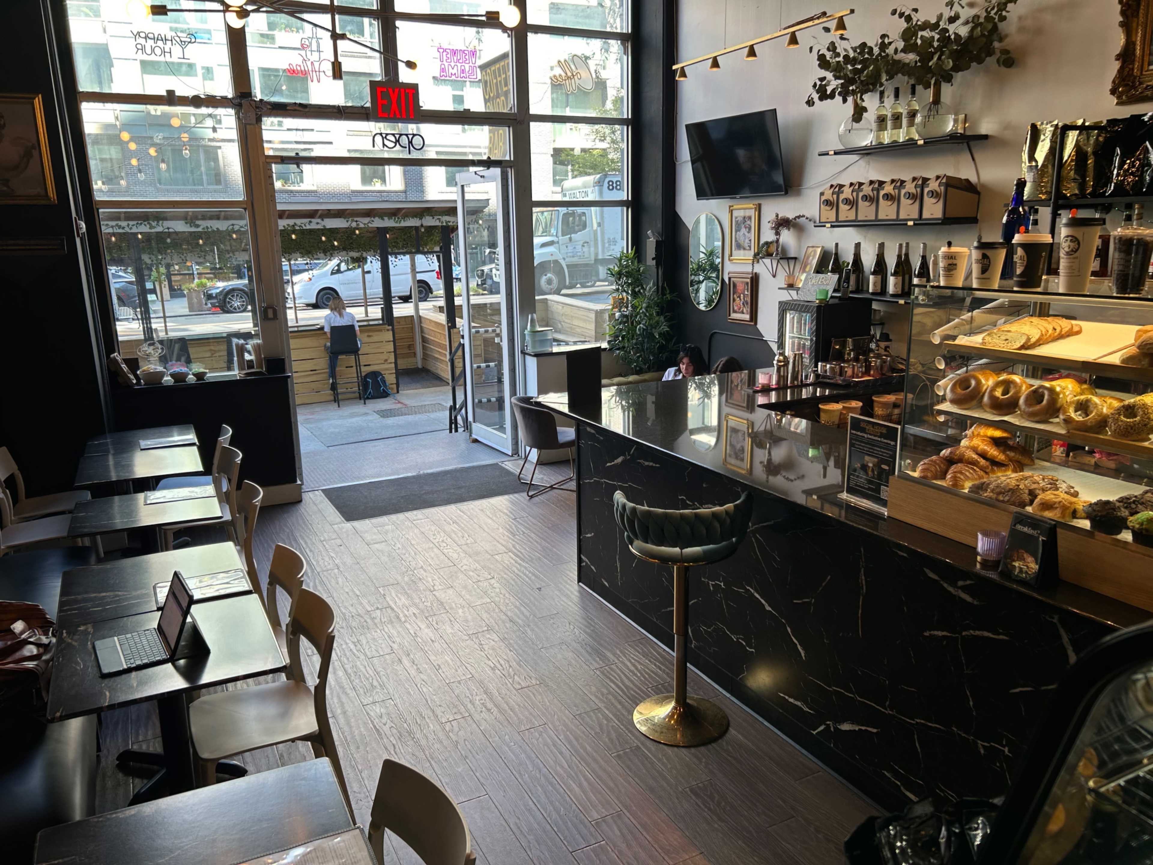The image shows a coffee shop interior with a counter displaying baked goods, rows of empty tables, and a large entrance framed by glass doors.