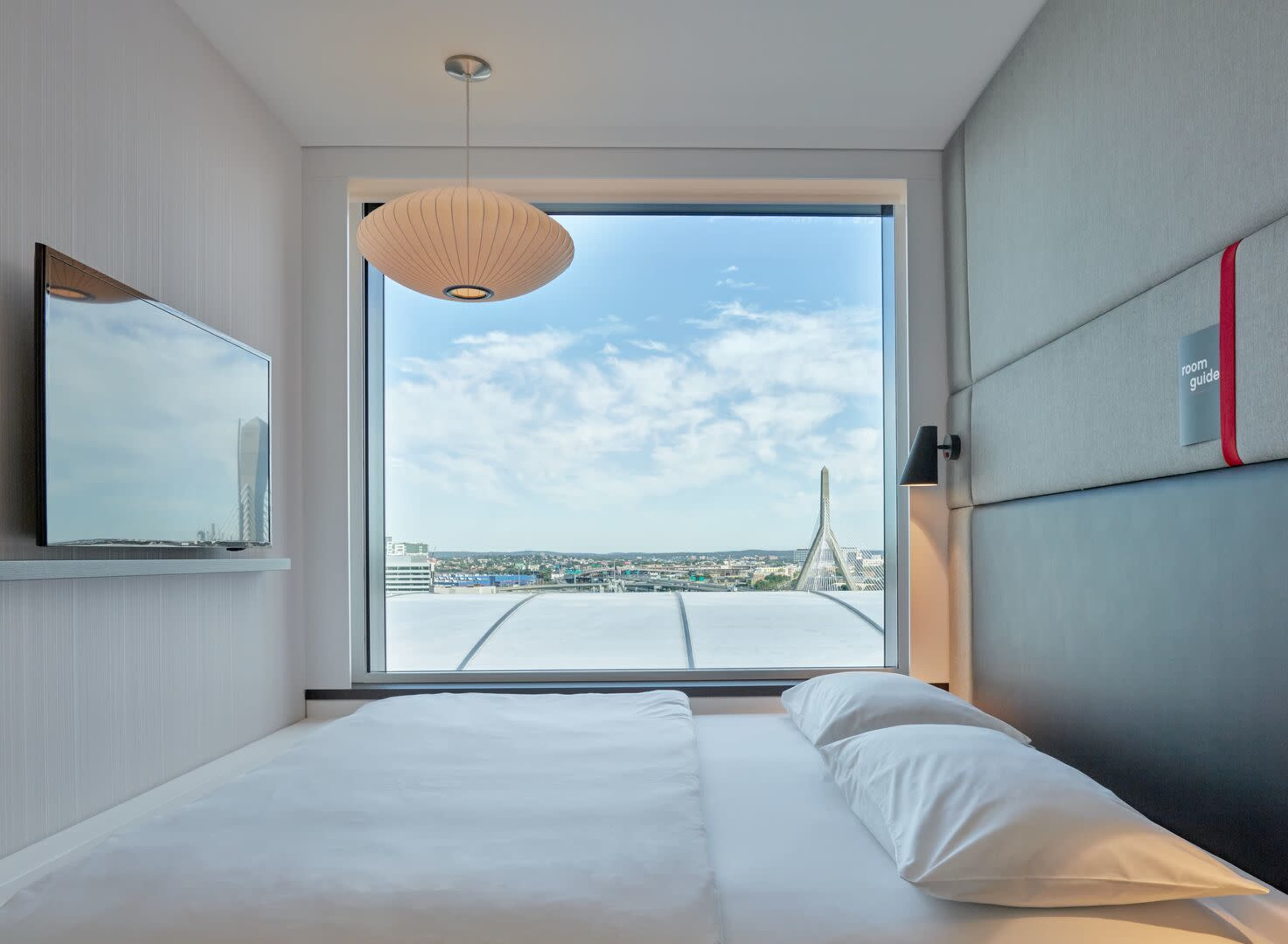A modern hotel room with a large window offering a view of a cityscape and a distinctive bridge.