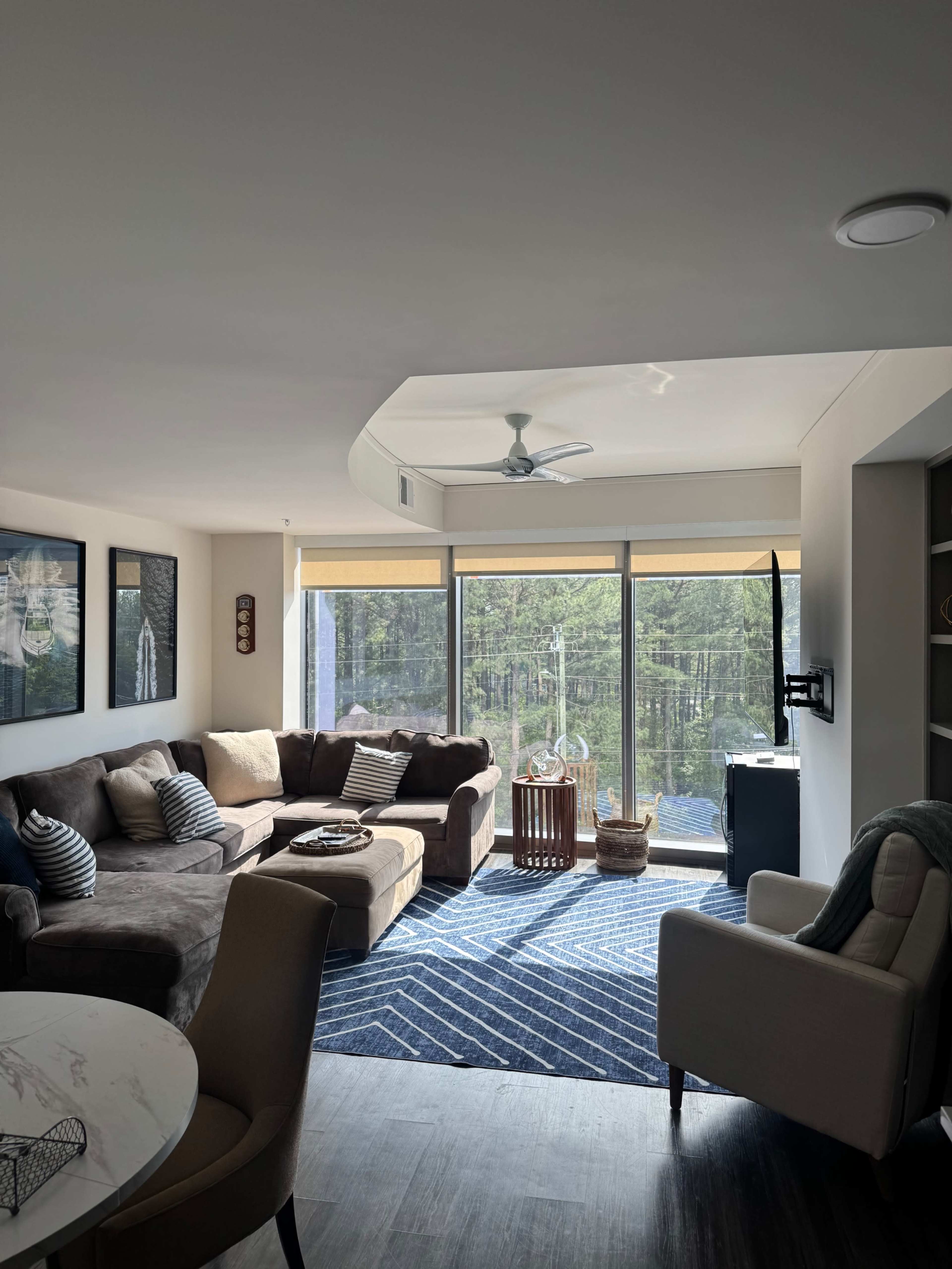 The image shows a modern living room with a brown sectional sofa, a circular dining table, and large windows overlooking a wooded view.