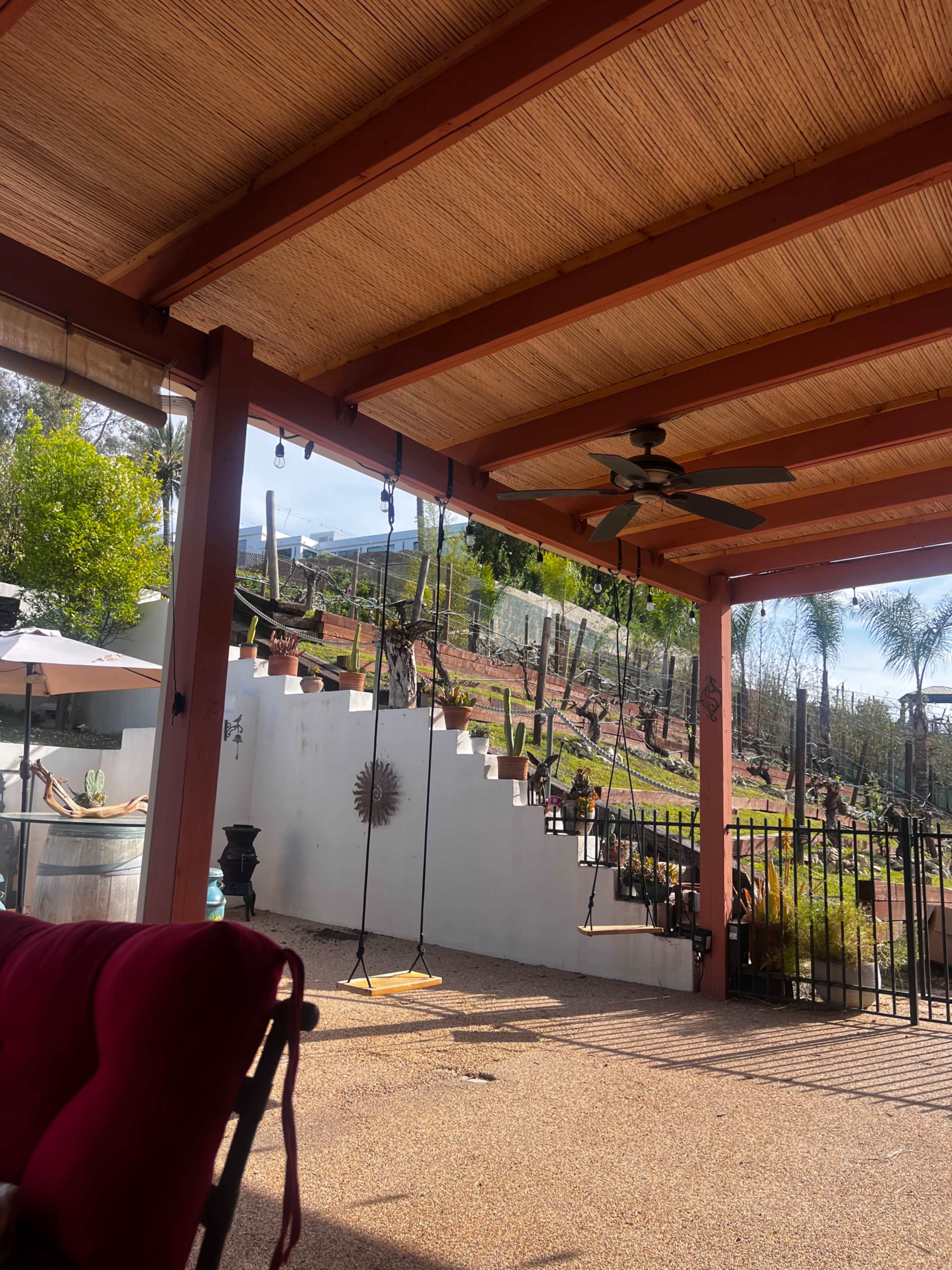 The image shows a shaded patio area with a swing, overlooking a hillside garden featuring steps and palm trees.