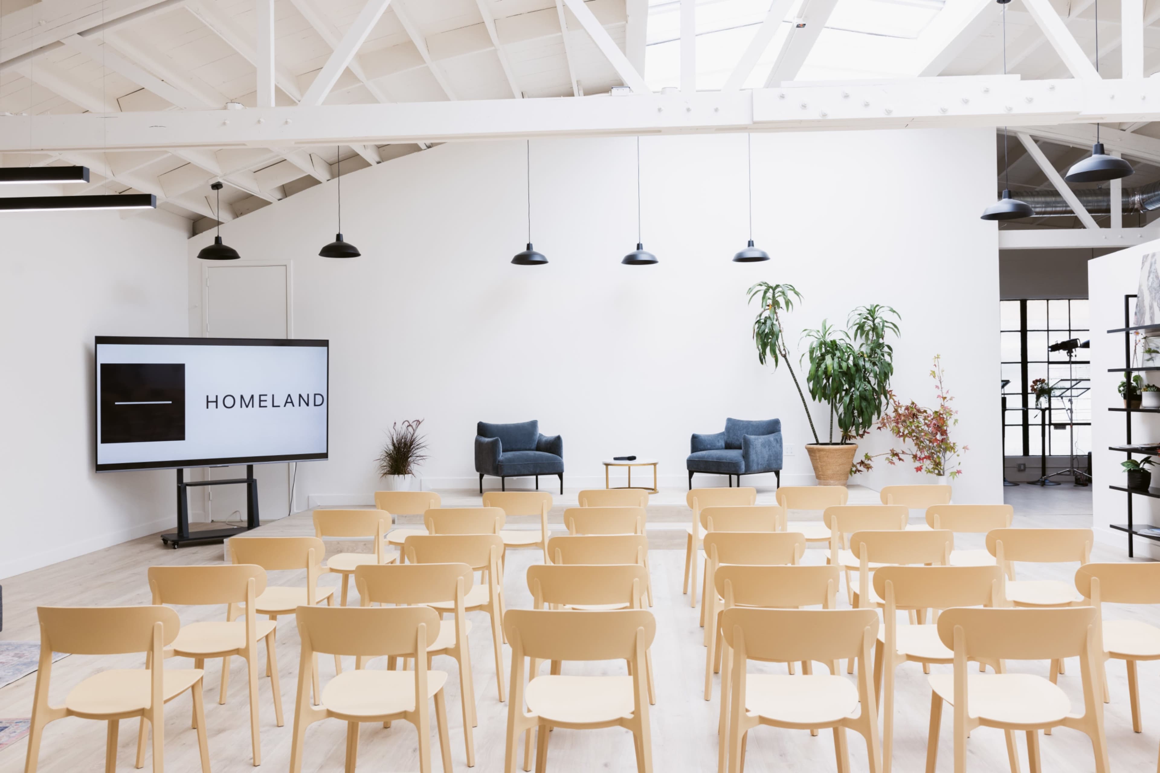 The image shows a spacious, well-lit room set up for a presentation, featuring rows of beige chairs facing a large screen displaying the word "HOMELAND," with two blue armchairs on a small stage.