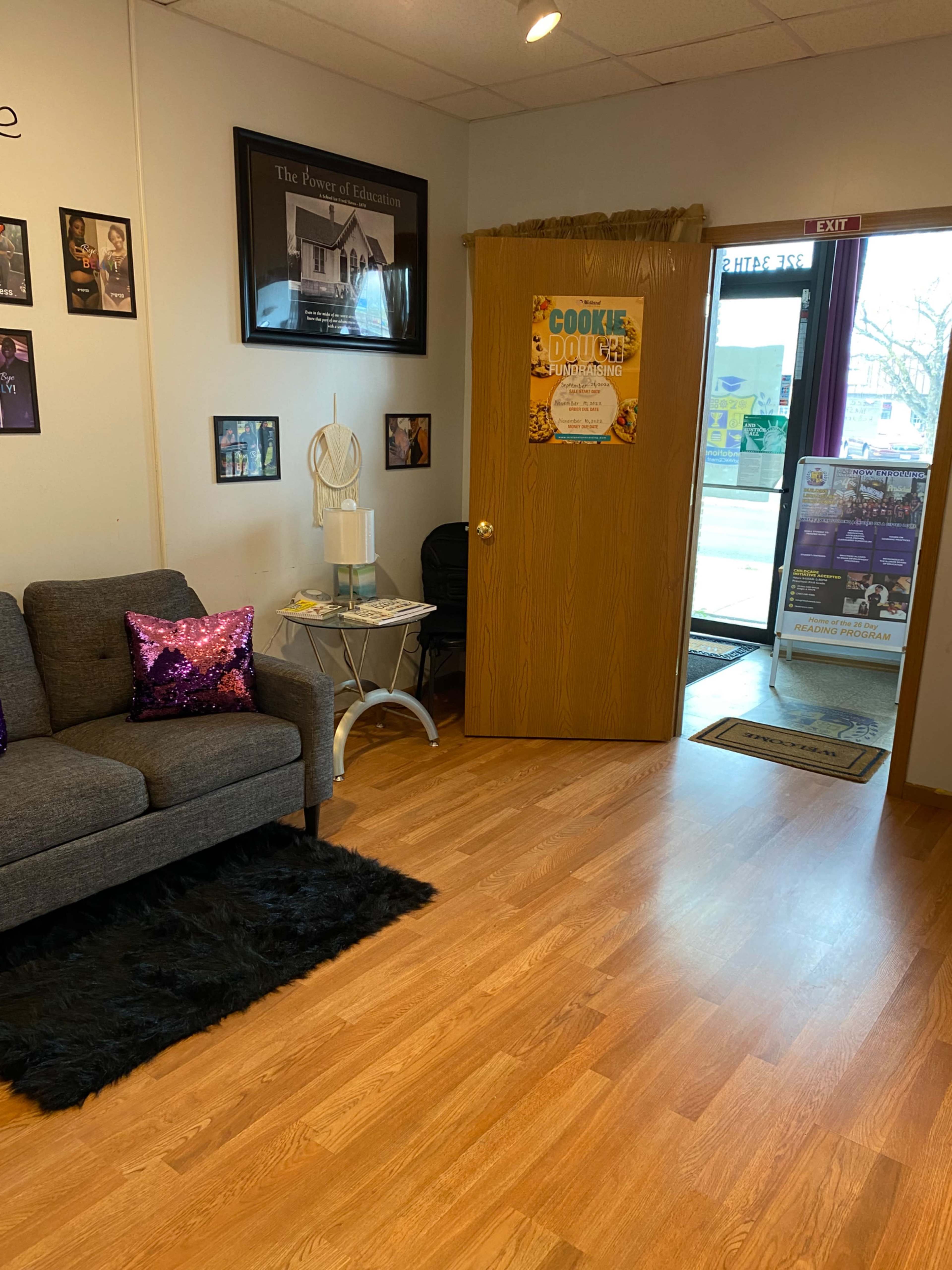 A cozy waiting area with a gray couch, a decorative rug, and a door leading to another room, alongside a bulletin board and posters on the walls.