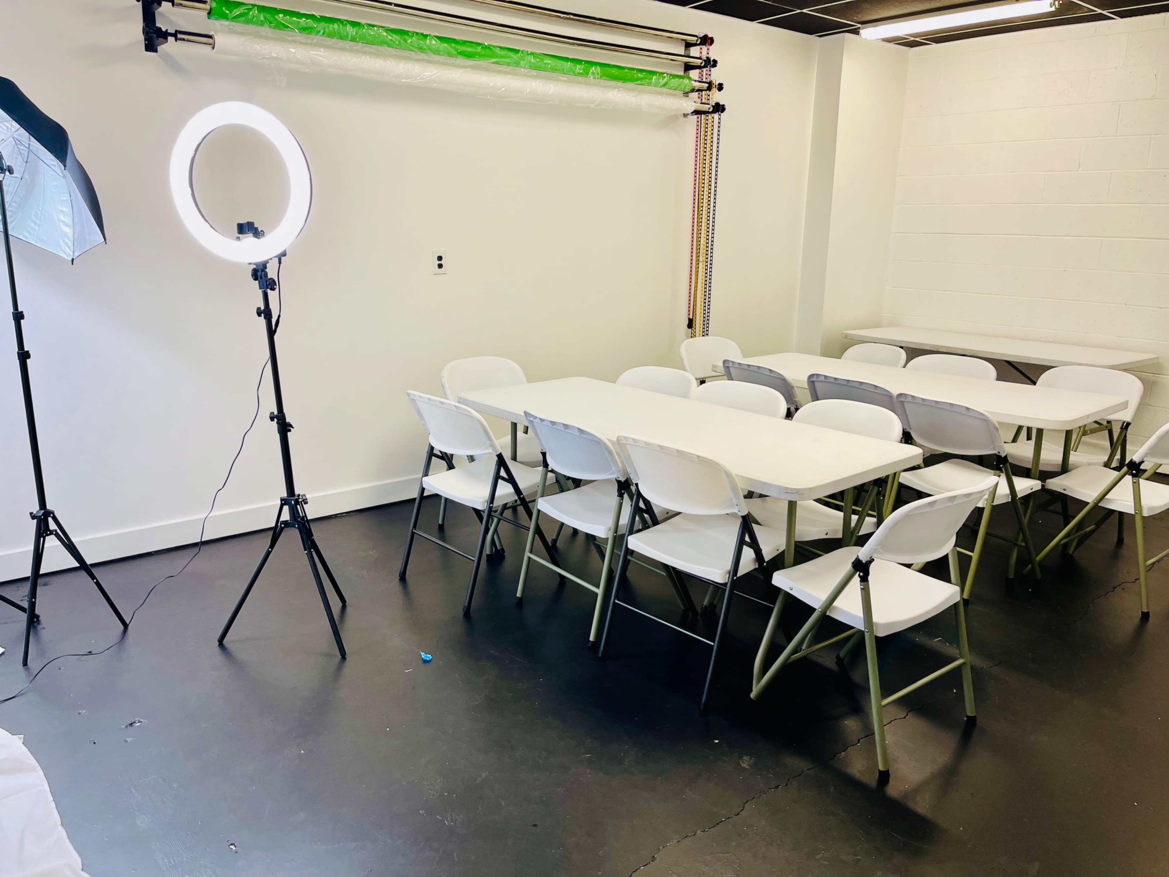 A plain room setup with a long table surrounded by chairs, a ring light on a stand, and a backdrop frame with a green screen.