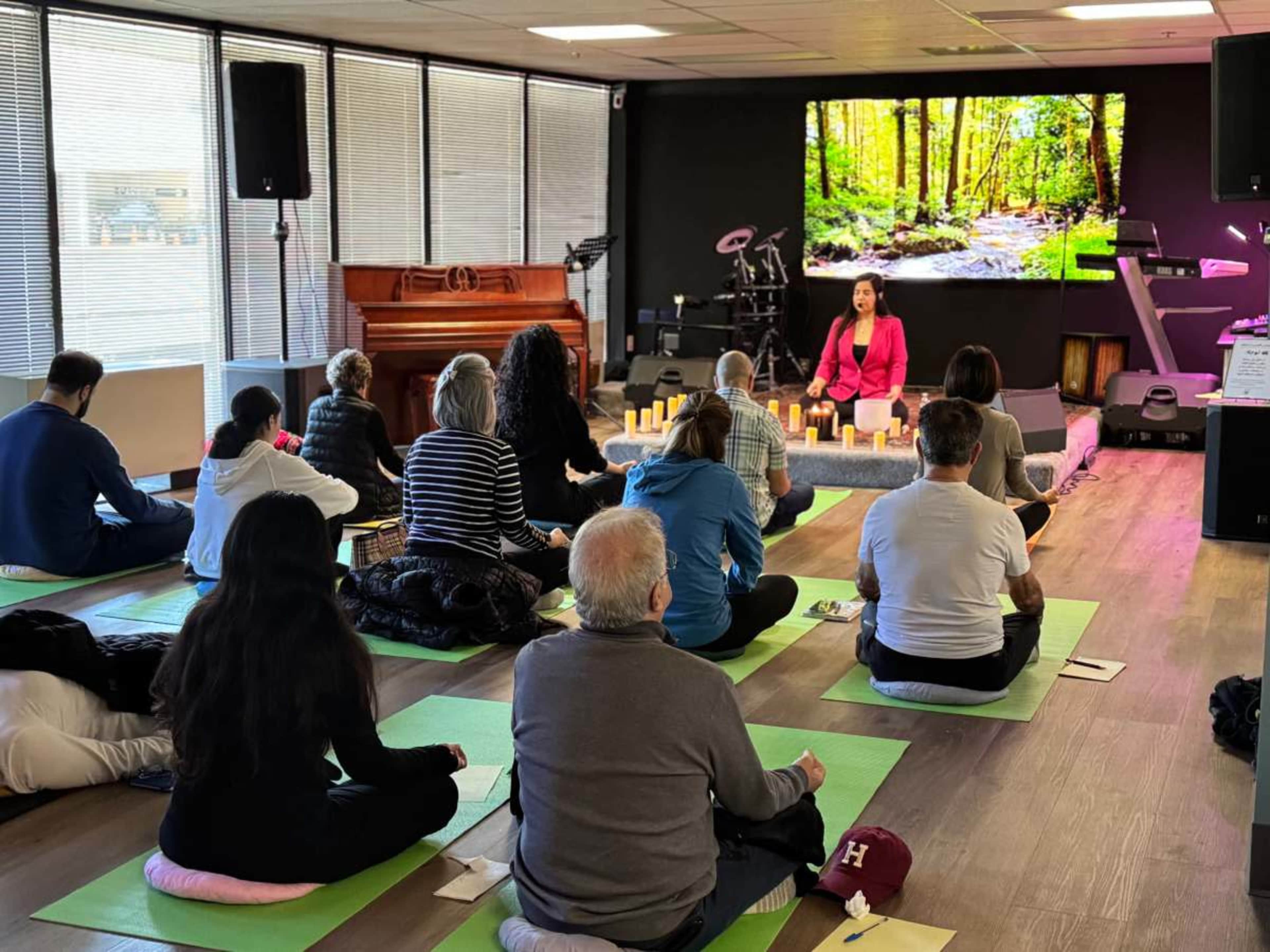 A group of people sits in a yoga studio with mats, facing an instructor who is seated in front of a large screen displaying a forest scene.
