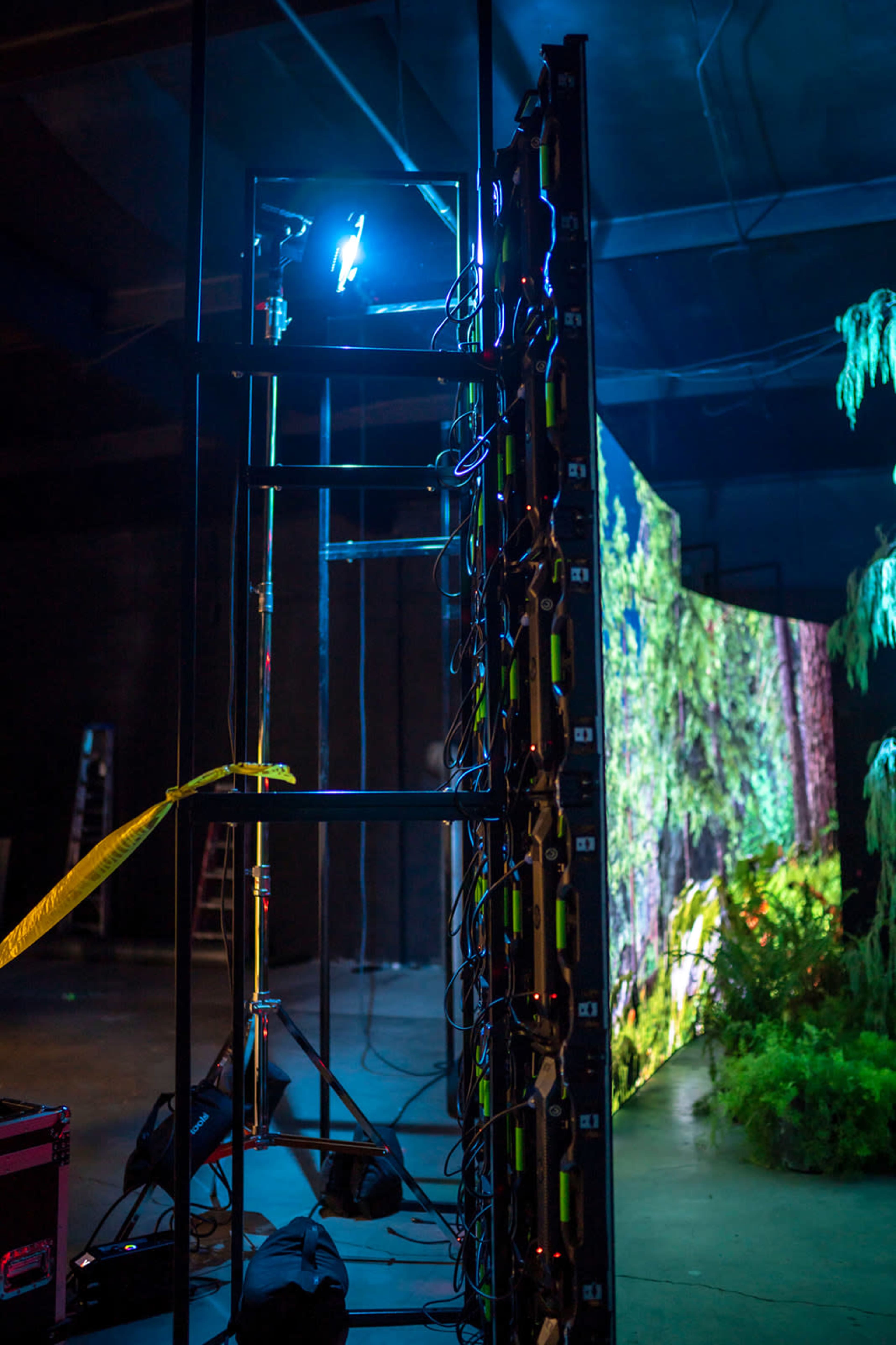 A tall scaffolding structure supports lighting equipment beside a large, illuminated backdrop displaying a nature scene in a dimly lit studio.