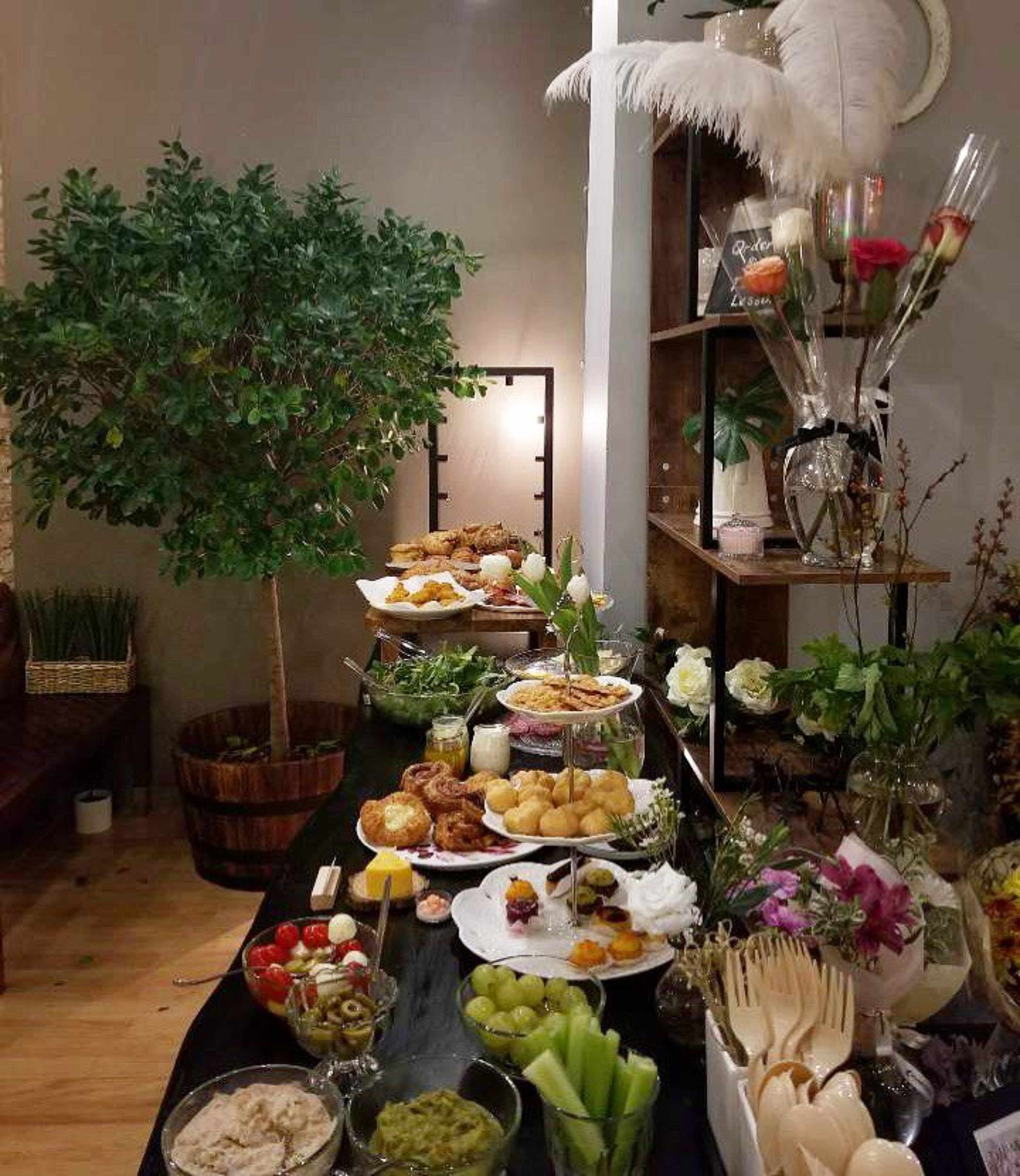 A table is set with various food platters, including fruits, pastries, and dips, alongside decorative plants and arrangements.