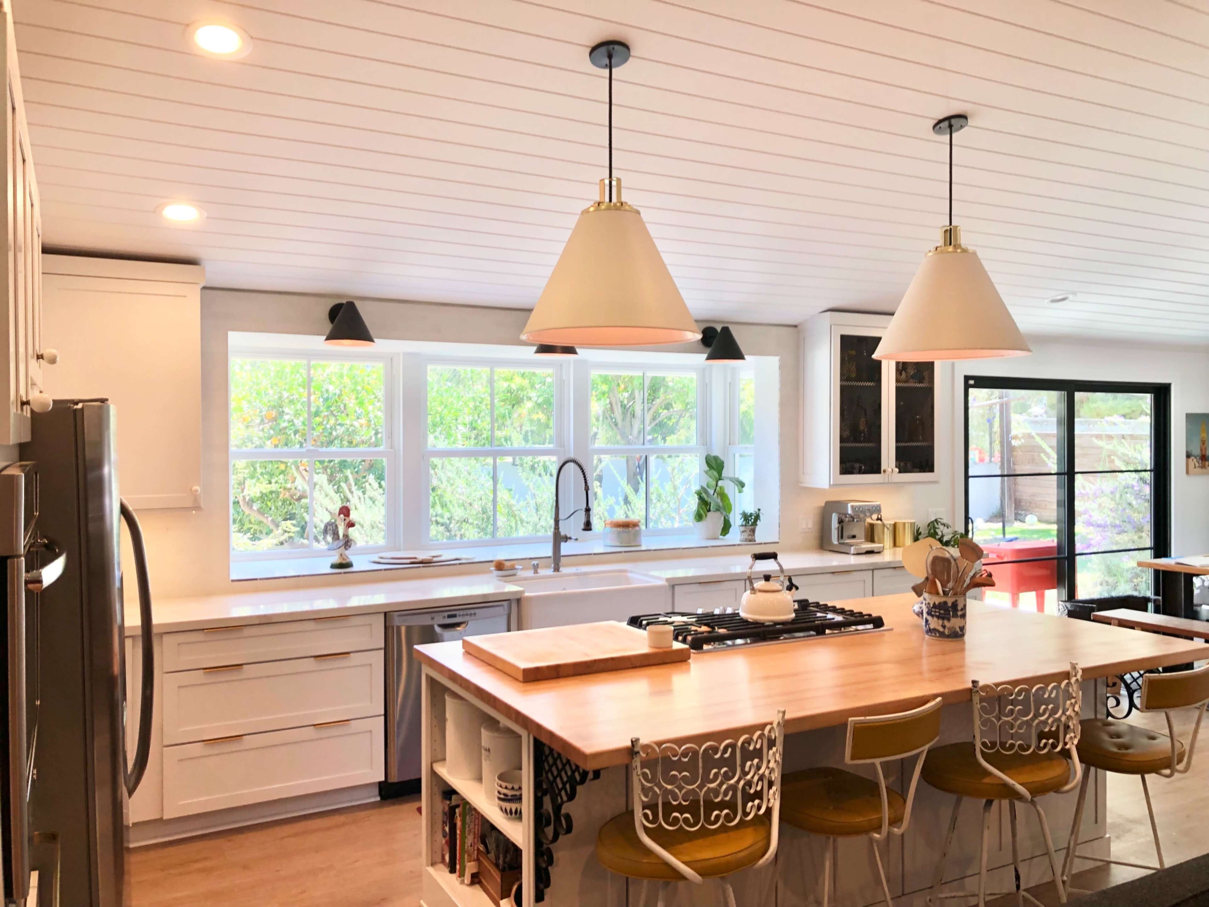 A bright kitchen with a central island, equipped with a gas cooktop and surrounded by bar stools, large windows allowing natural light, and various kitchen appliances.