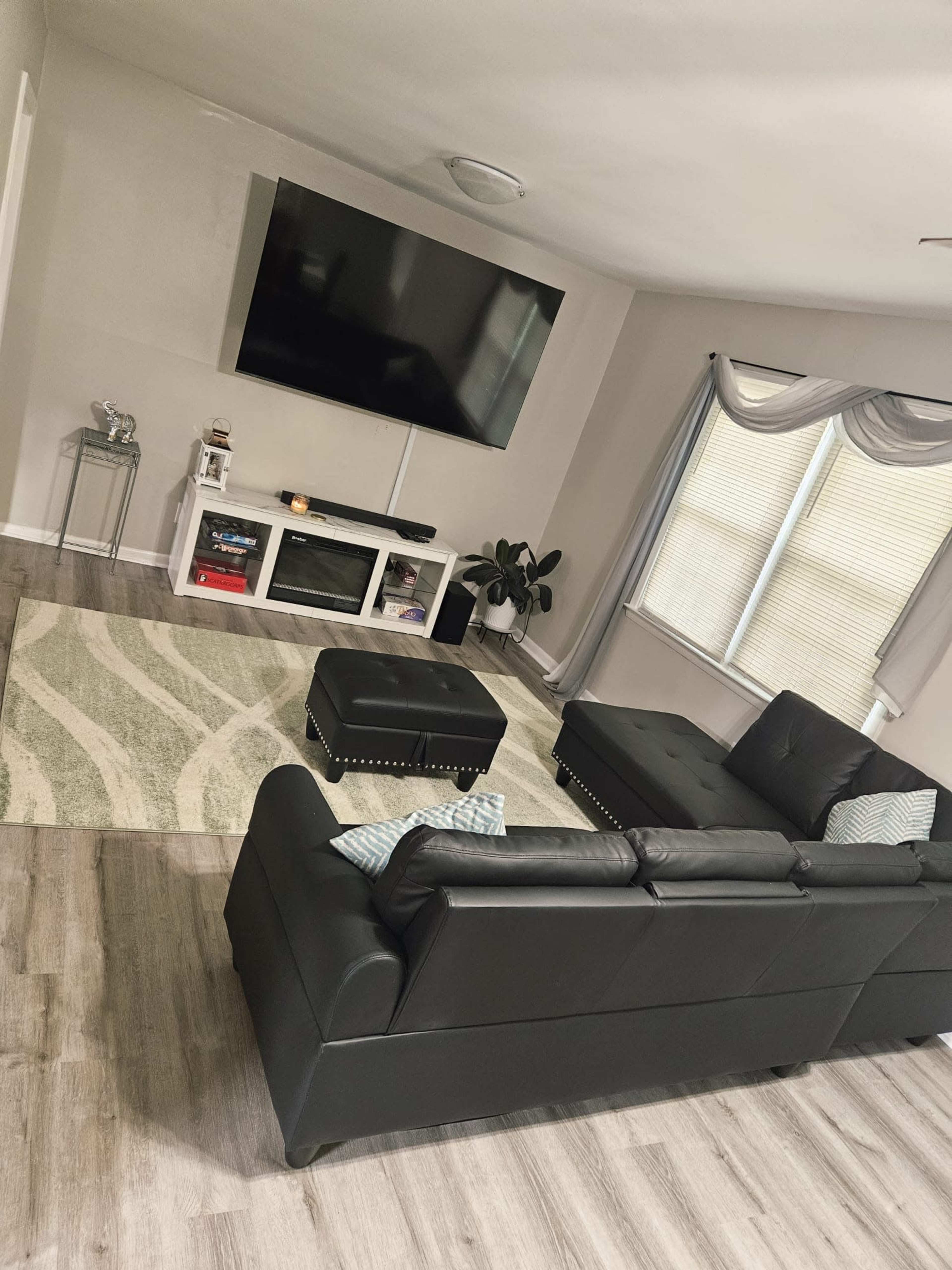 A black leather sectional couch faces a large wall-mounted television above a white media console in a well-lit living room with light-colored walls and a patterned area rug.