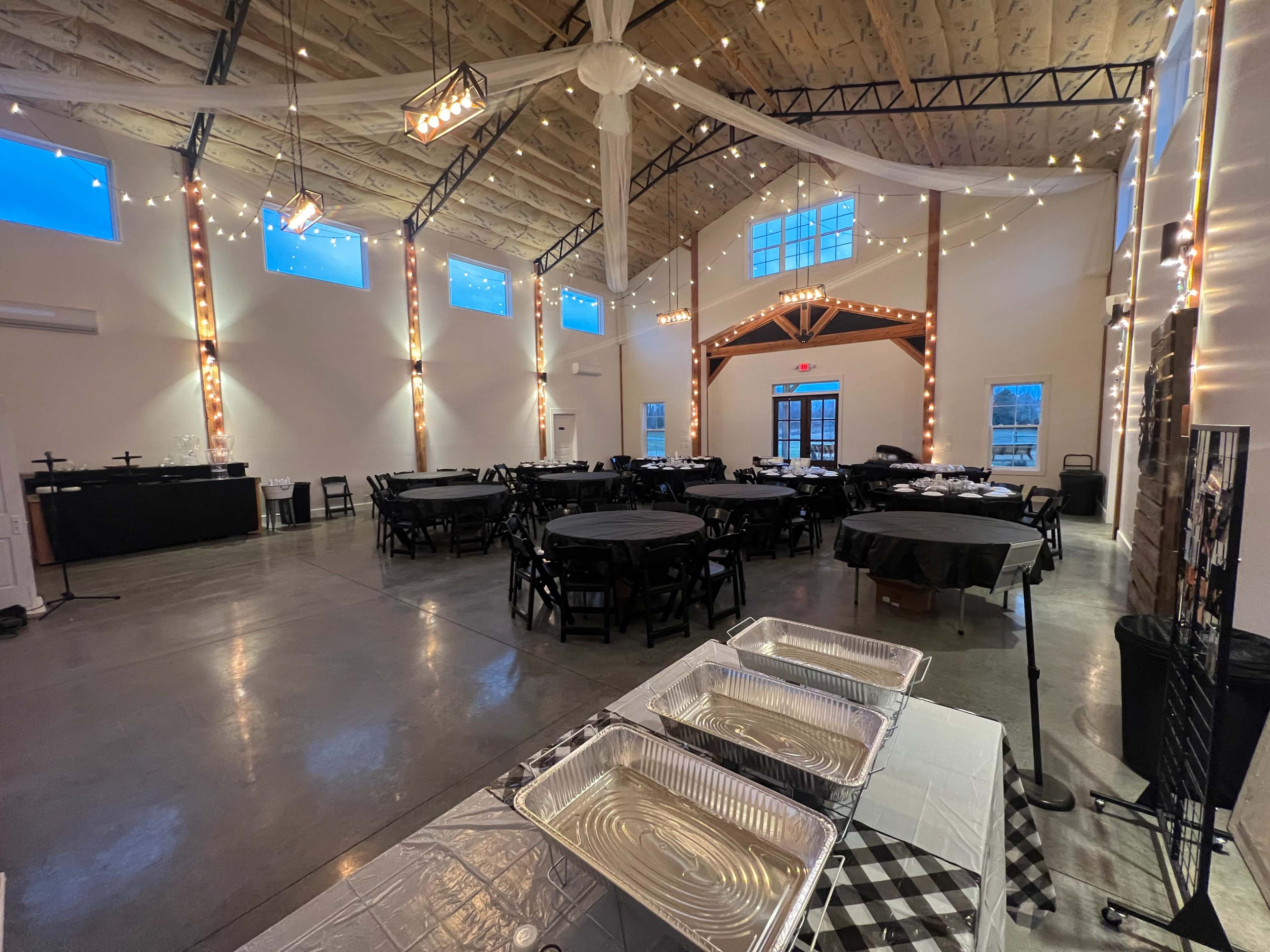 A spacious event hall with round black tables set for dining, string lights overhead, and a service table with aluminum trays in the foreground.