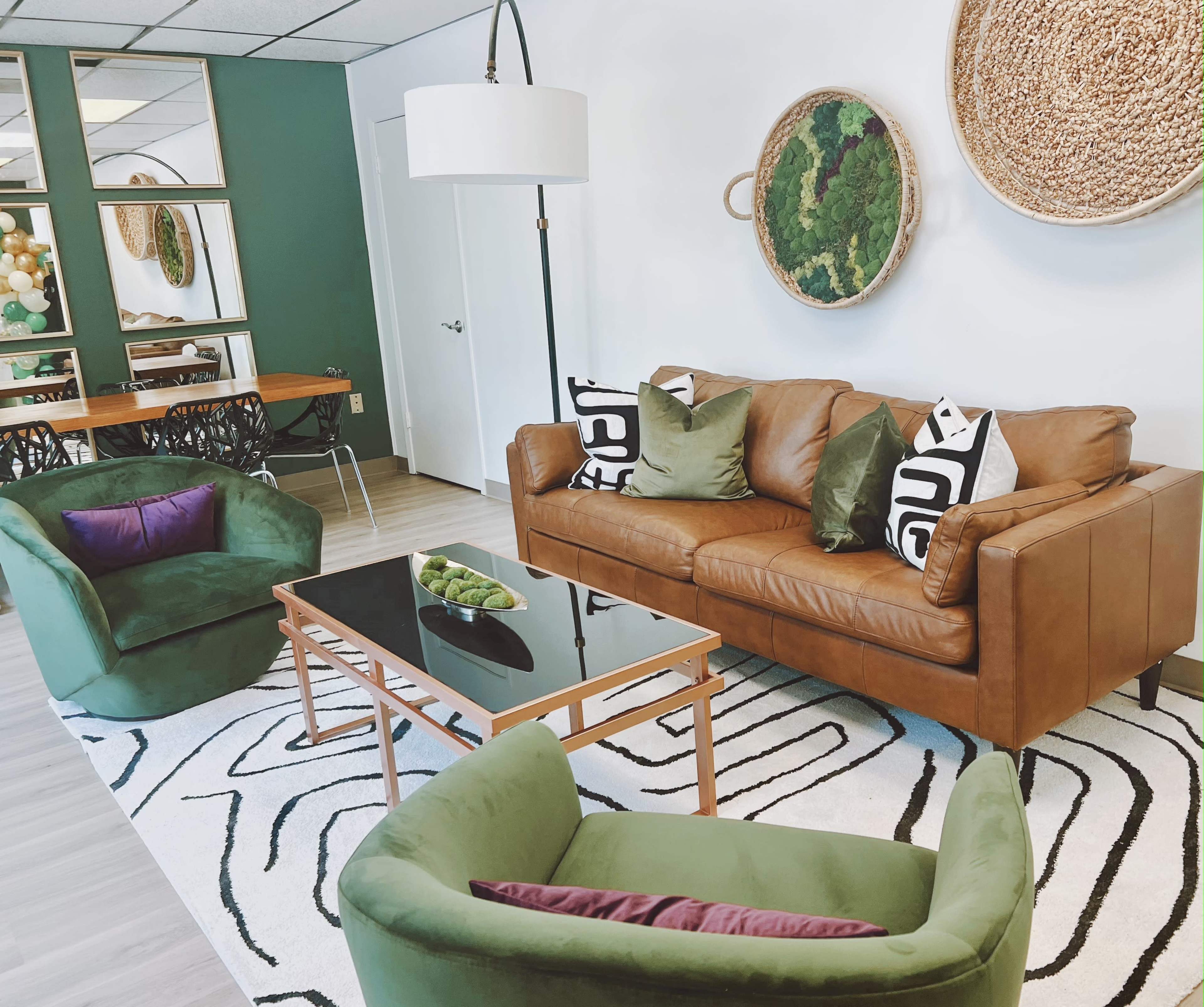 The image shows a stylish living room with a brown leather sofa, two green chairs, a glass coffee table, and wall decor featuring woven baskets.