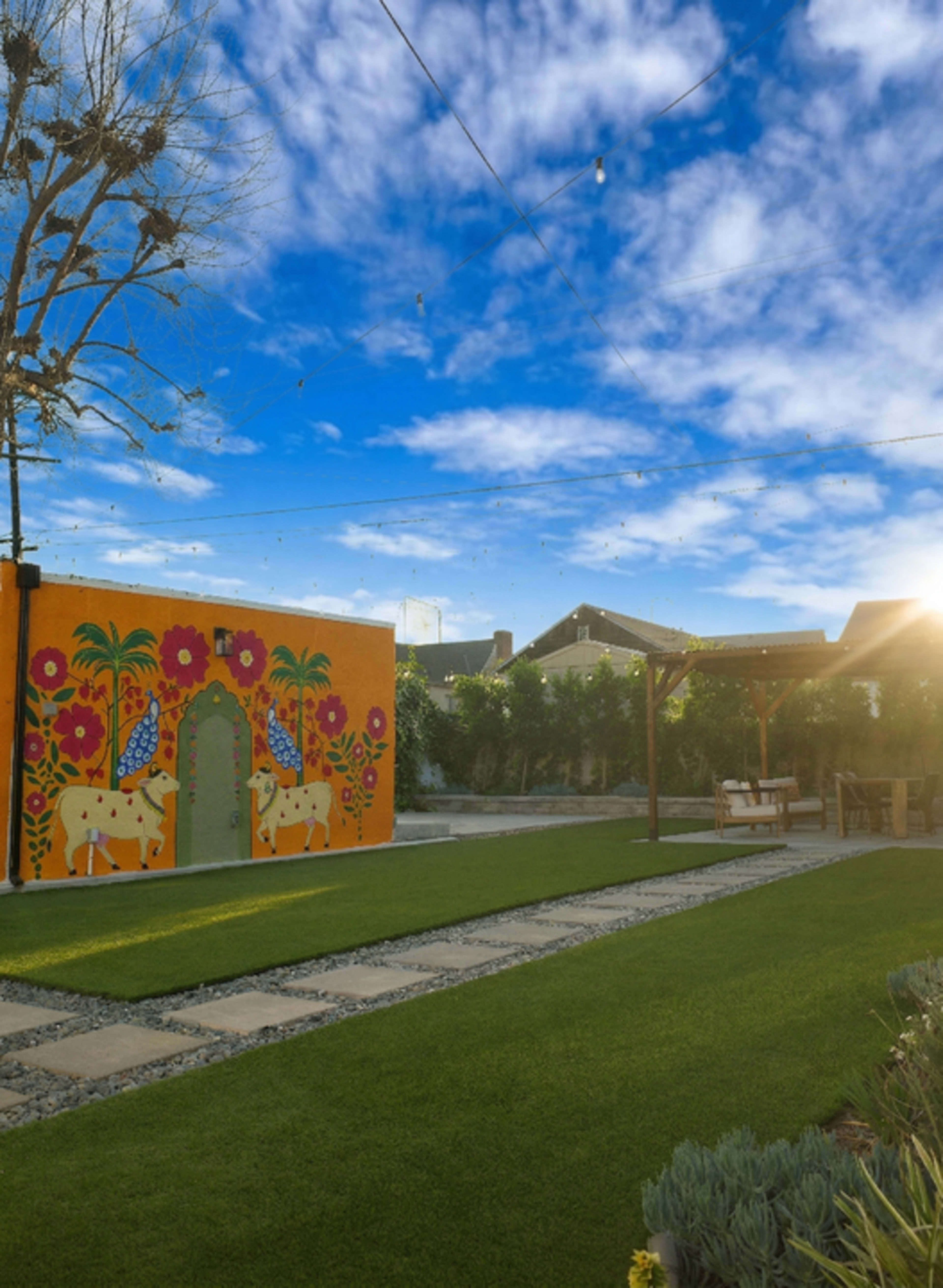 A colorful mural featuring cows and vibrant plants adorns a wall in a landscaped backyard with green grass, stone pathways, and a shaded seating area.